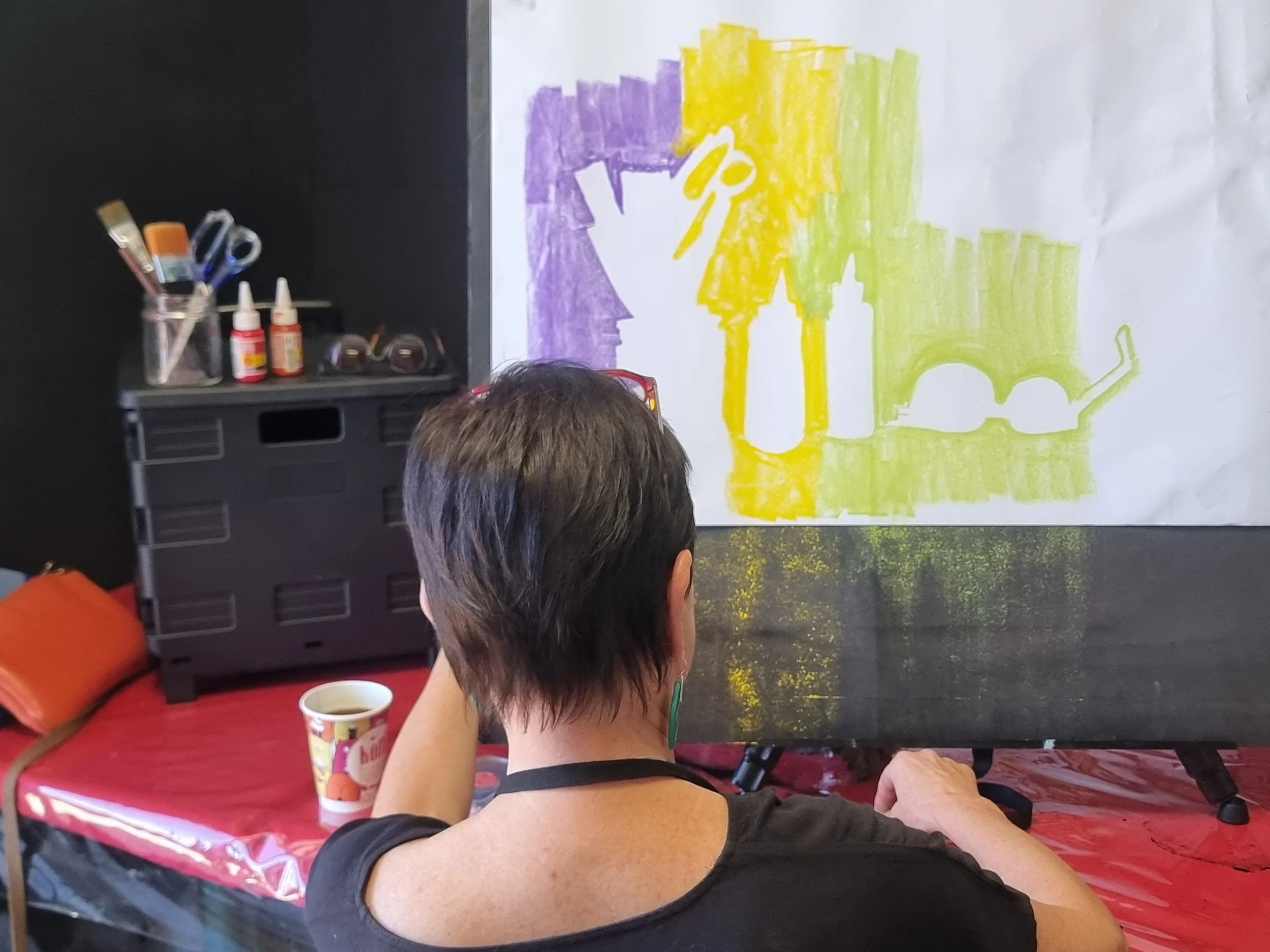 Woman with short dark hair sitting at a red-covered table, watching a colorful drawing of glass bottles, a pair of glasses, and a rectangular shape on a large piece of paper. The drawing features yellow, purple, and green sections. Beginner art class