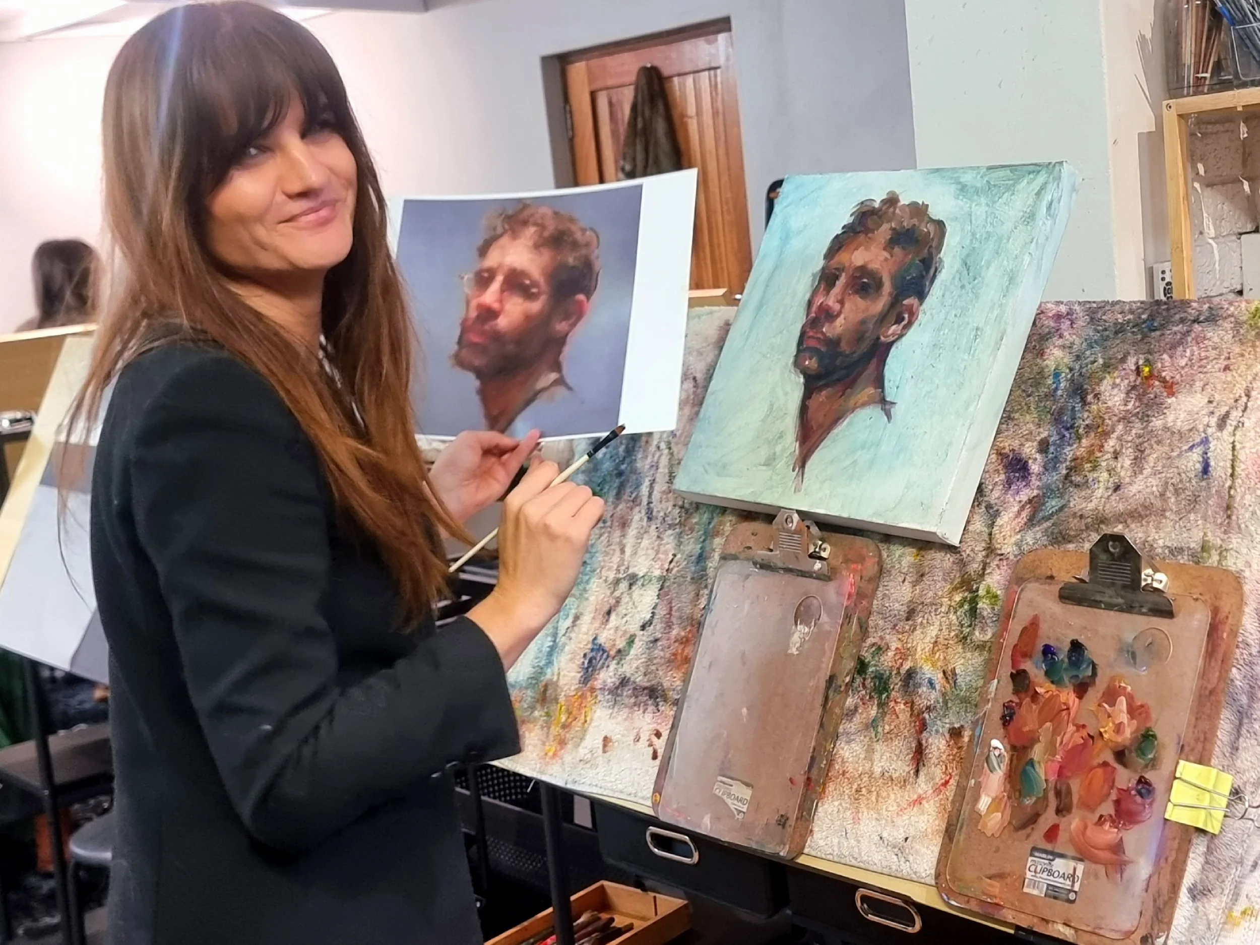 A woman with long brown hair and a black blazer is smiling at the camera while holding a paintbrush in her right hand. She is in an art studio, working on a portrait of a man with short, curly hair and a beard. Her painting is in progress on a canvas