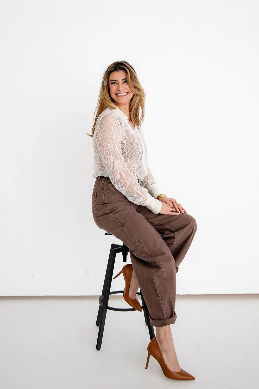 A young woman sitting on a metal chair with her legs spread, wearing ripped jeans and a pink pleated blouse, resting her chin on her hand, smiling at the camera, against a plain white background.