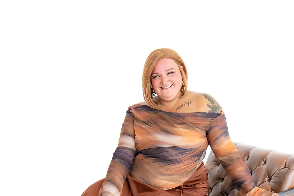 Woman with pink, wavy, shoulder-length hair smiling, wearing a black leather jacket draped over her shoulders, a black and white striped sleeveless top, and beige pants, standing against a plain white wall.