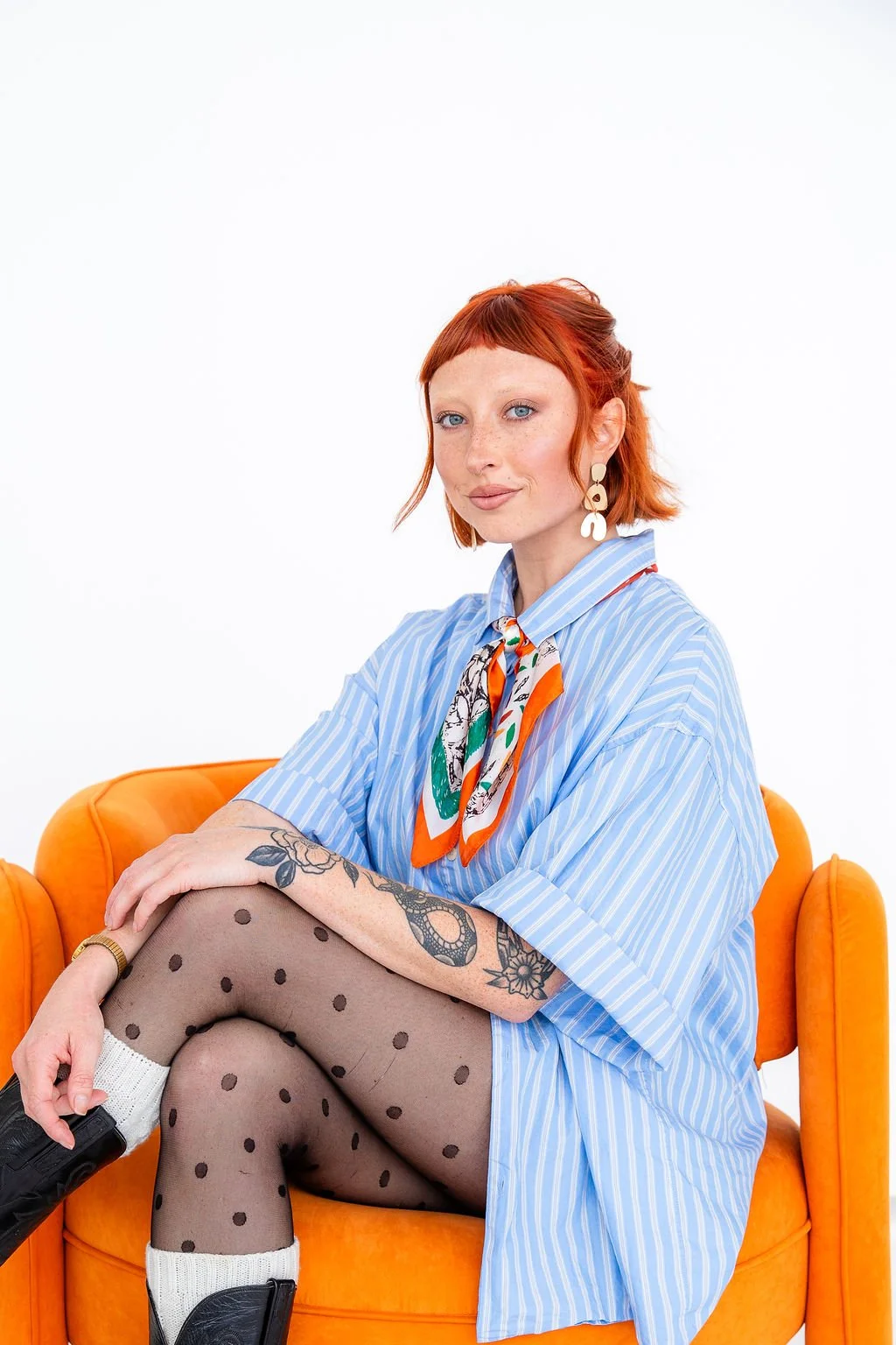 A woman with short, blond hair, wearing a black sleeveless top, a brown bandana around her neck, and gold hoop earrings, smiling with a tattoo of a woman, a man, a dog, and a flower on her arm.