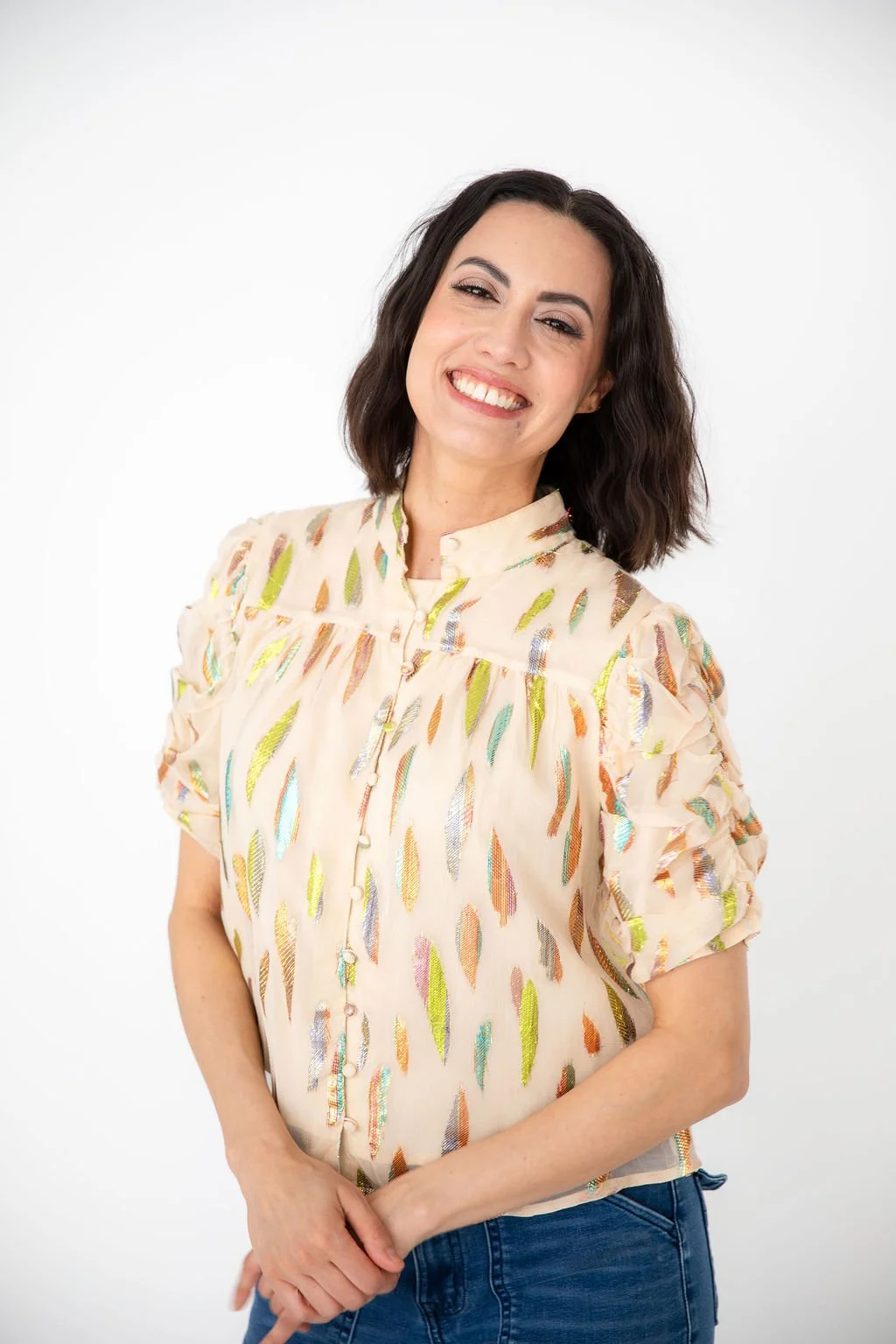 A woman with dark wavy hair smiling, wearing a white embroidered blouse with ruffled sleeves and a maroon leather skirt, standing against a black background.