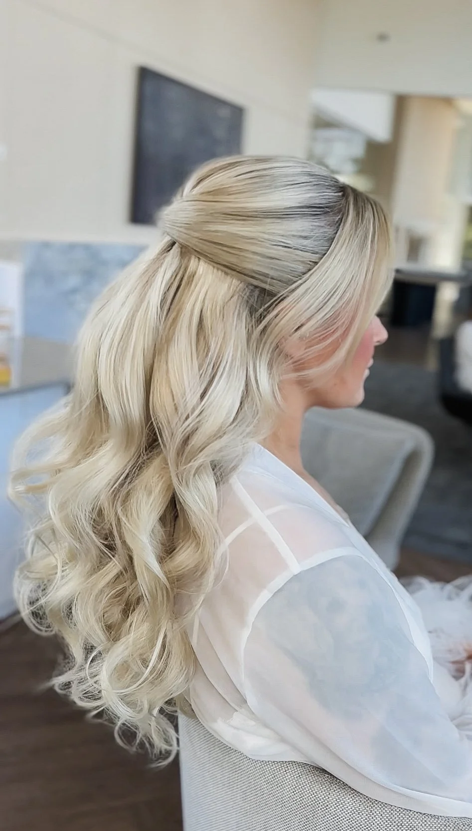 Side view of a woman with long, wavy blonde hair styled with a half-updo, sitting indoors.