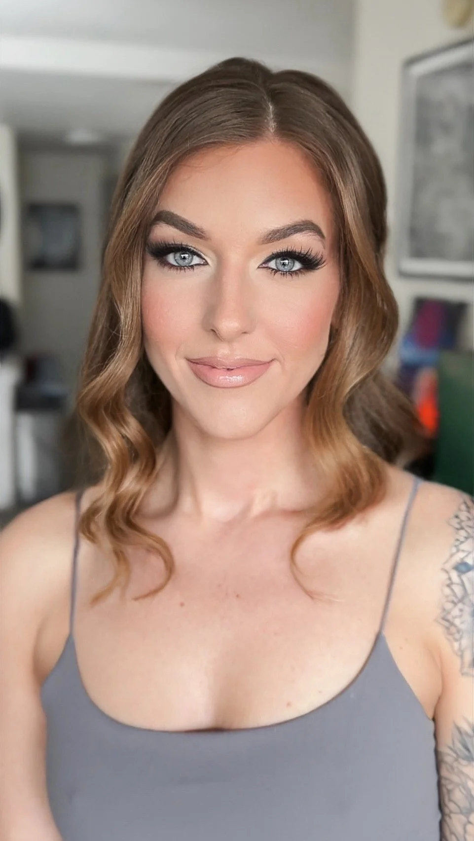 A woman with wavy, shoulder-length brown hair, blue eyes, and makeup, wearing a gray spaghetti-strap top. She has a tattoo on her left shoulder.