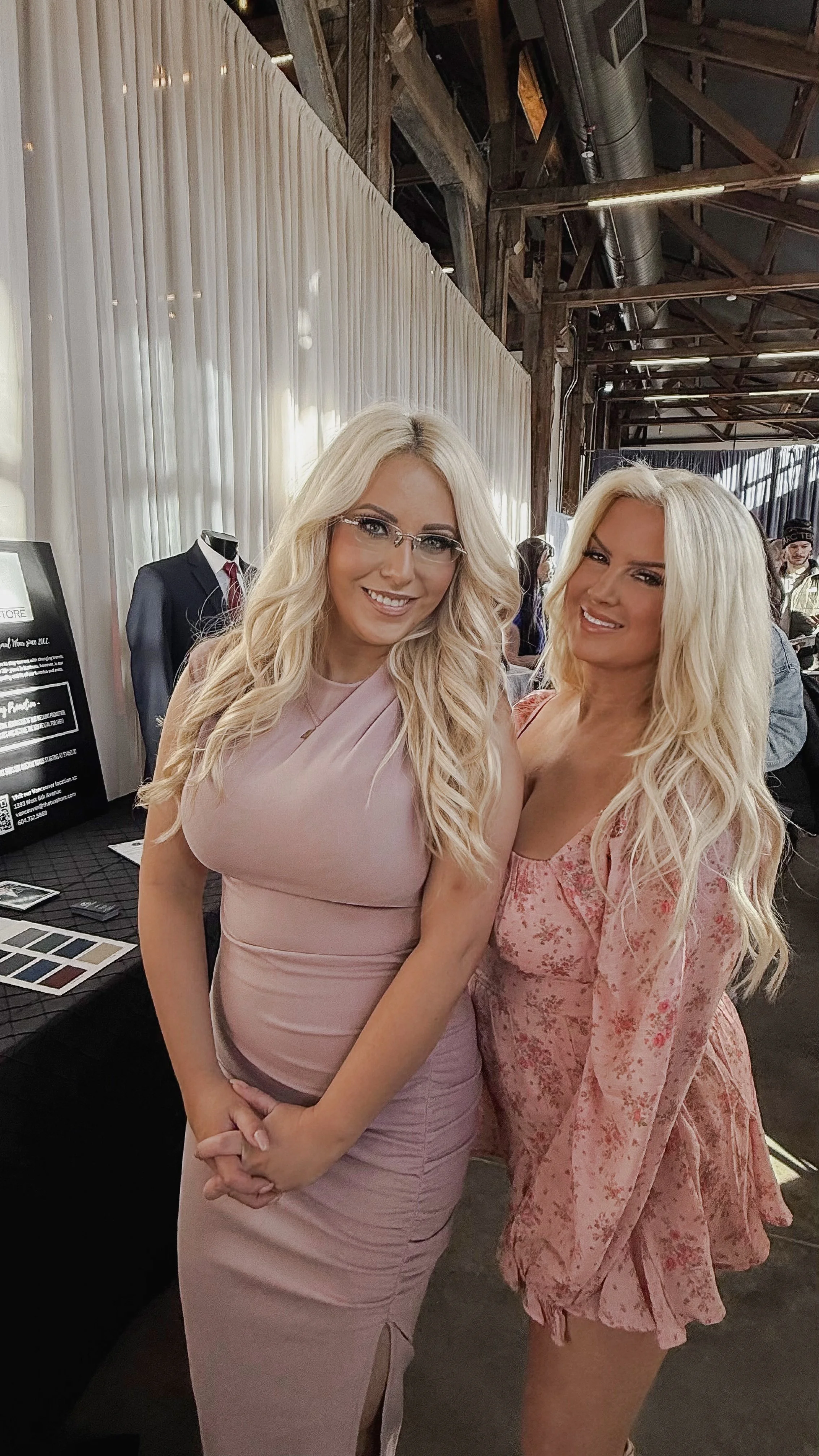 Two women with long blond hair, wearing pink dresses, are smiling and standing close together indoors at an event with wooden beams and white curtains.