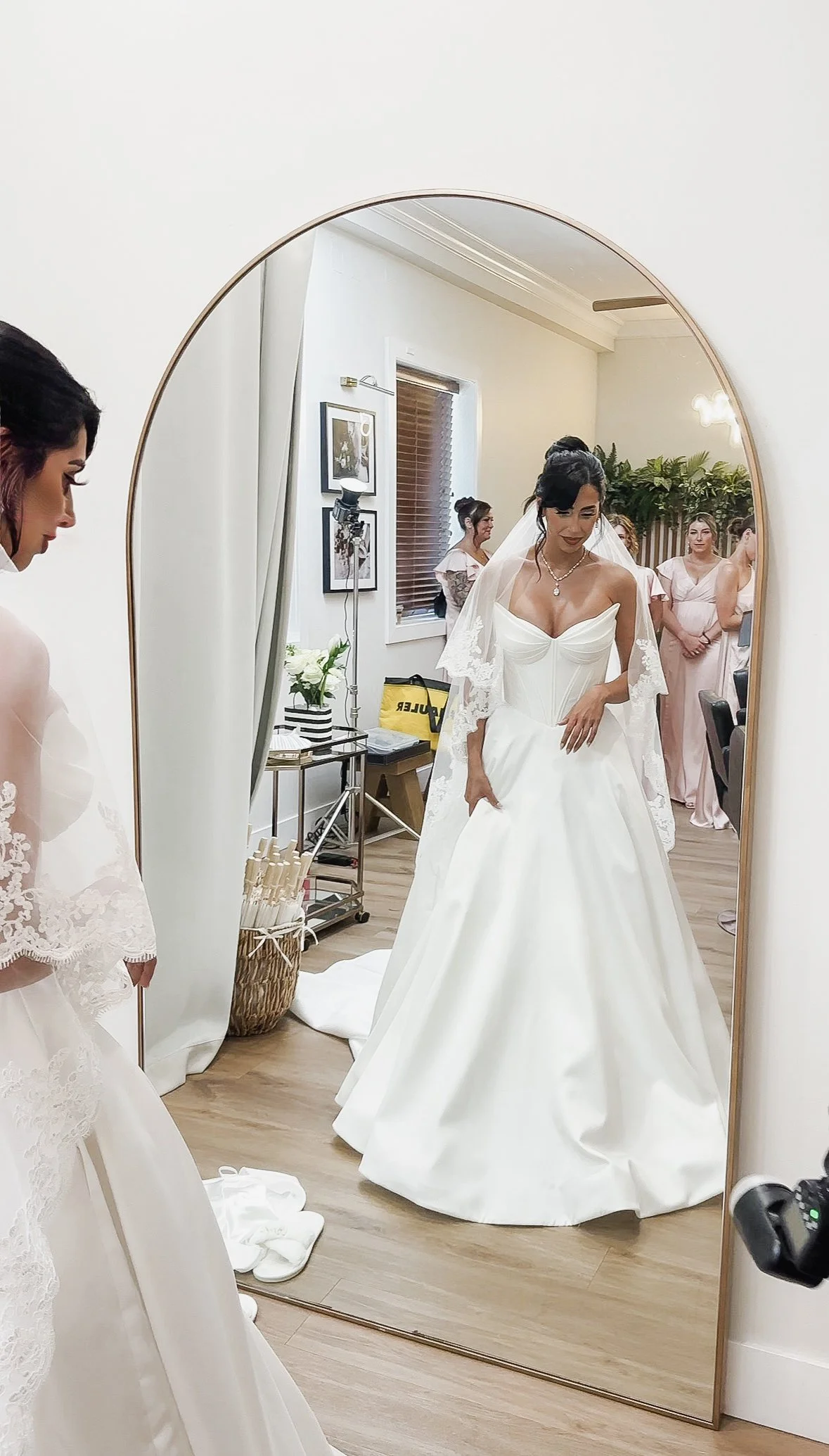 A bride trying on her wedding dress, looking in a full-length mirror, with her bridesmaids and wedding attendants in the background.