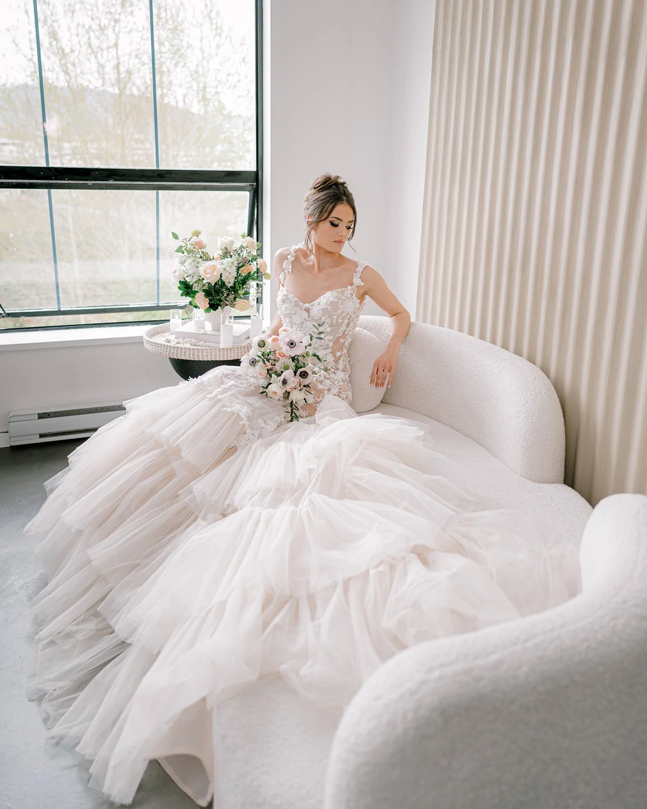 Bride in a white lace wedding gown sitting on a cream sofa, holding a bouquet of white and pink flowers, near a large window with greenery outside.