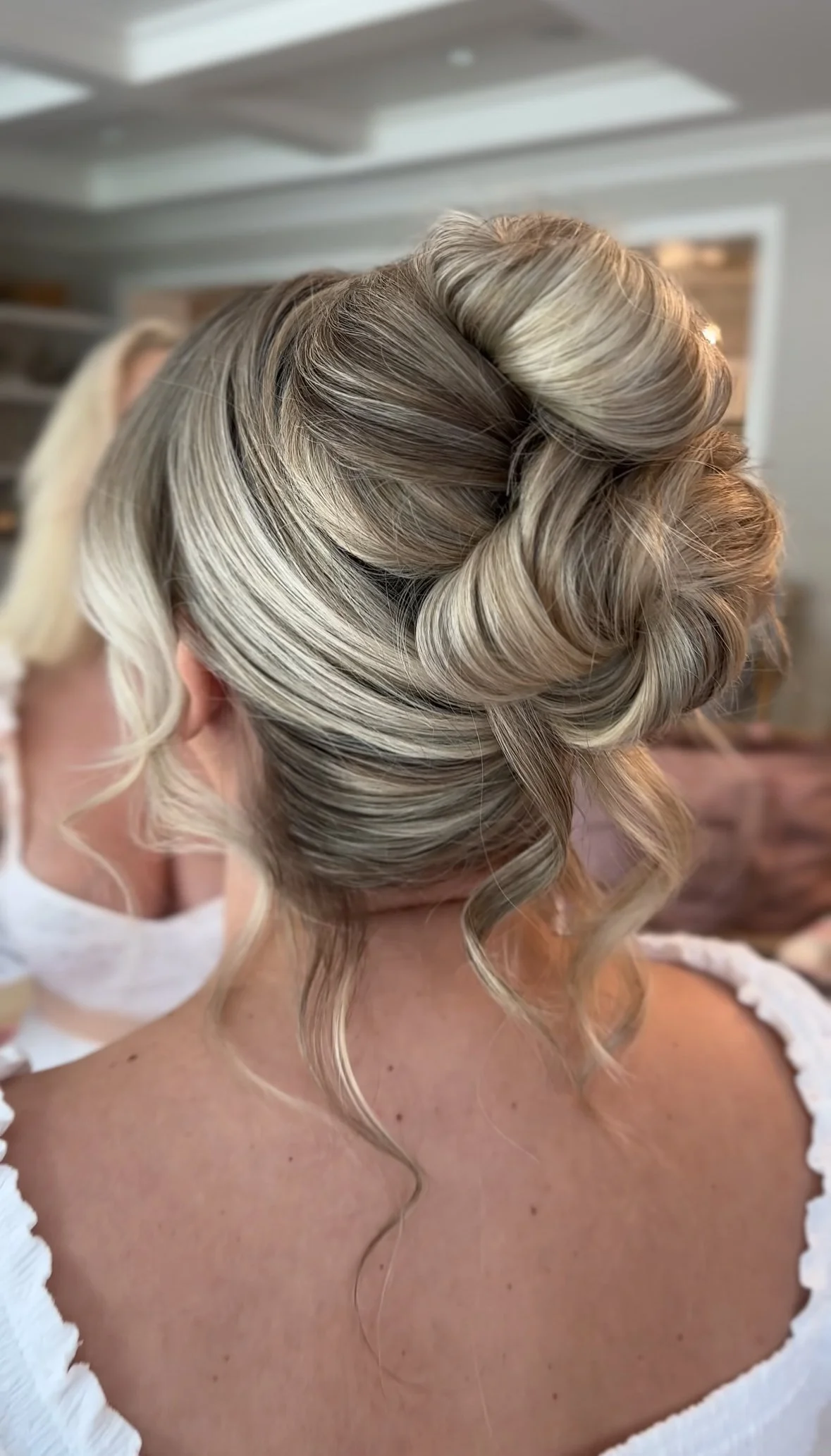 Up-close view of an elegant, intricately styled blonde updo with curls and twists, set against a blurred indoor background.