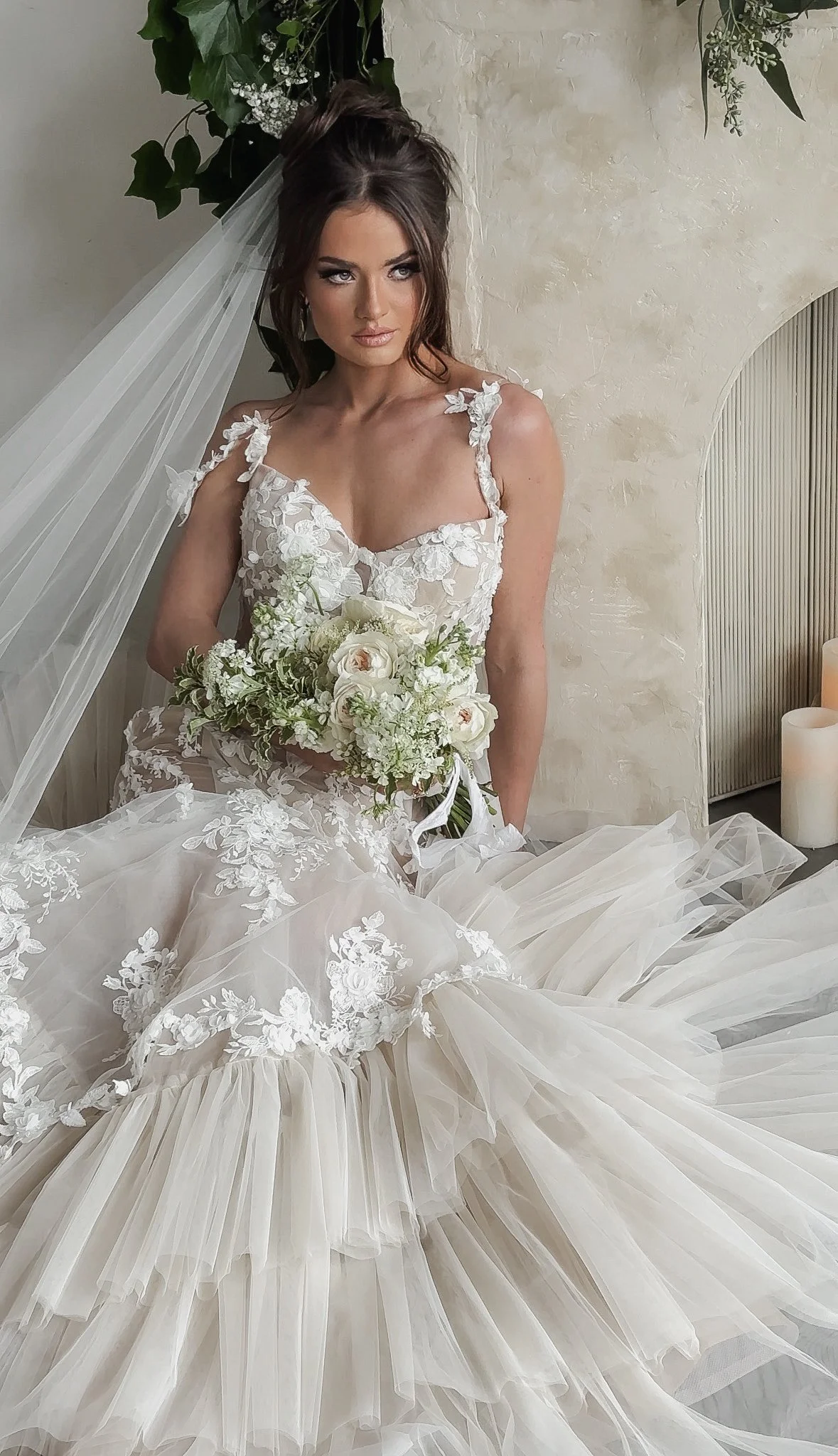 A bride sitting on a ledge, wearing a lace wedding gown with embroidered floral details, holding a bouquet of white flowers, with a curtain of candles and green plants in the background.