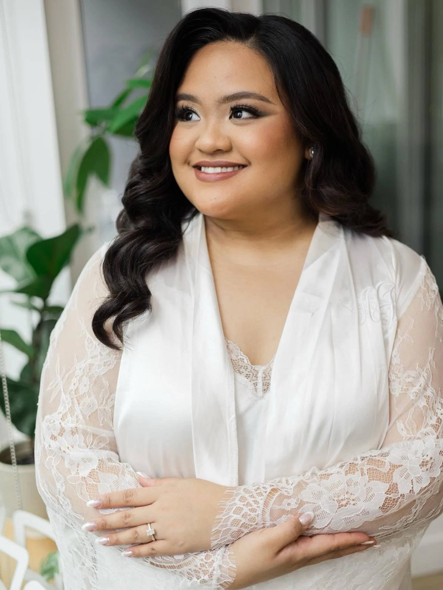 A woman with long black wavy hair and makeup, smiling, wearing a white lace robe, standing indoors near a window with green plants in the background.