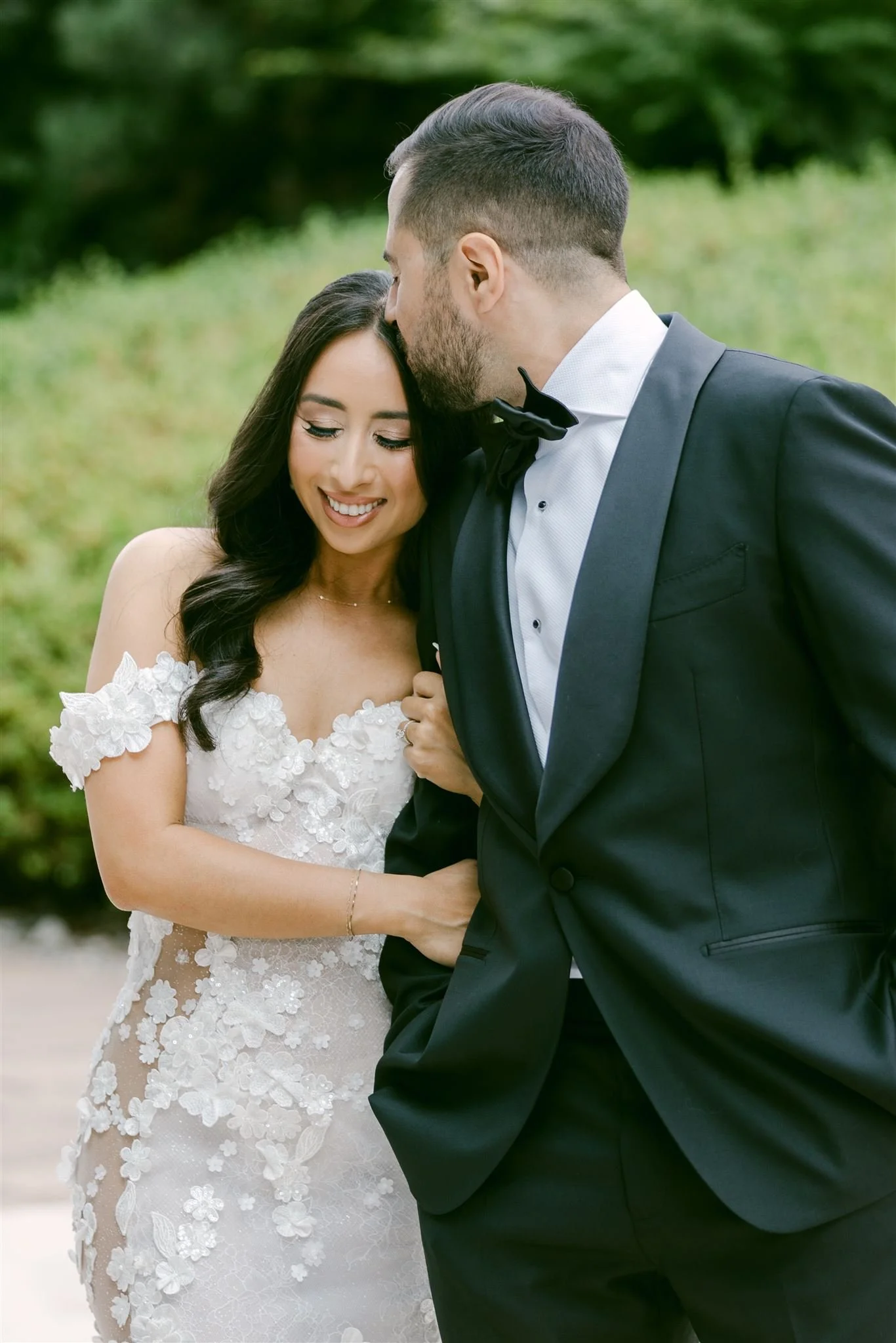 A bride and groom in wedding attire sharing an intimate moment outdoors, the groom kissing the bride on her forehead.