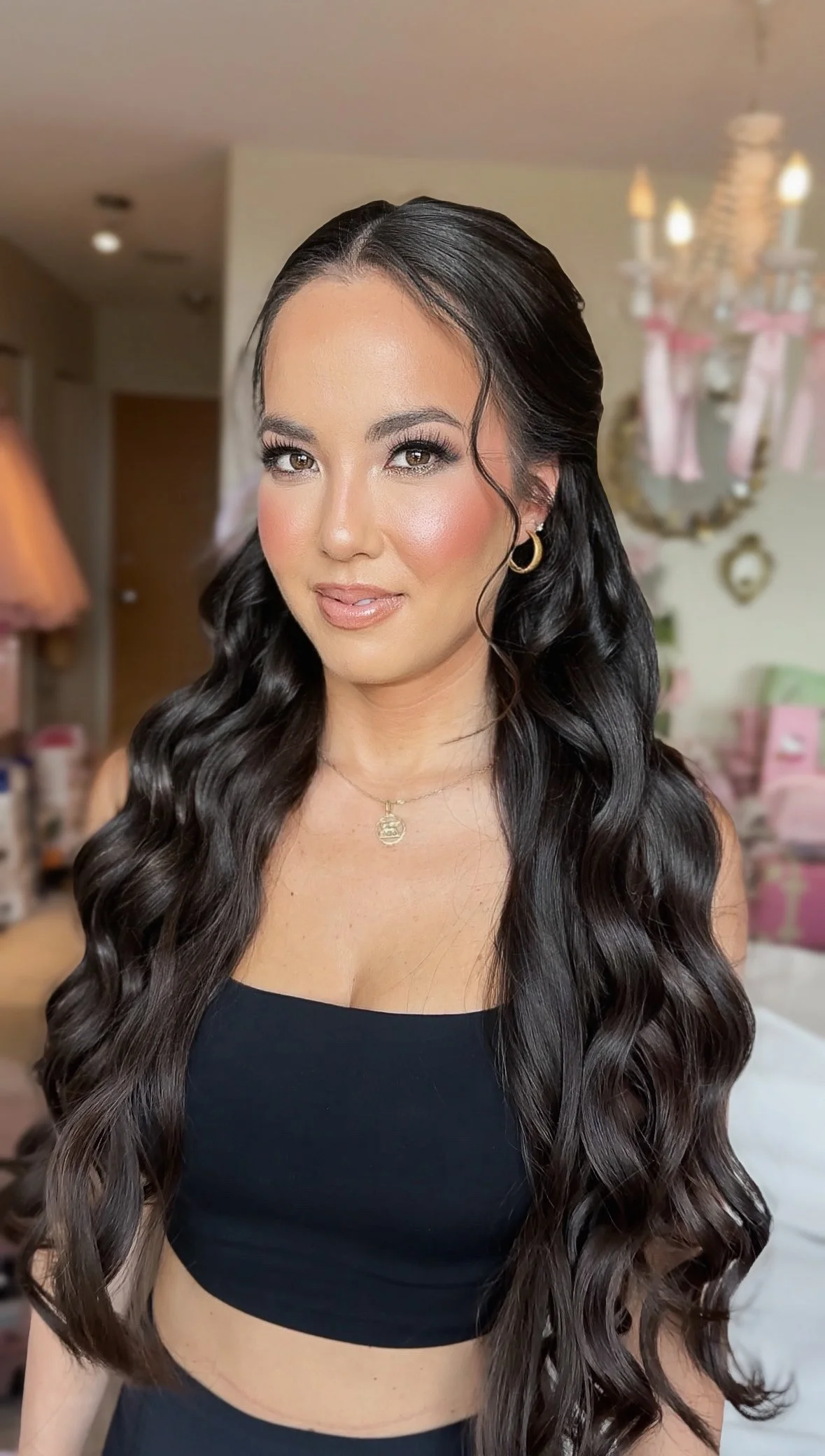 A woman with long, wavy black hair wearing a black top, gold hoop earrings, a gold necklace, and makeup, standing in a room with pastel decorations including a chandelier and pink accents.