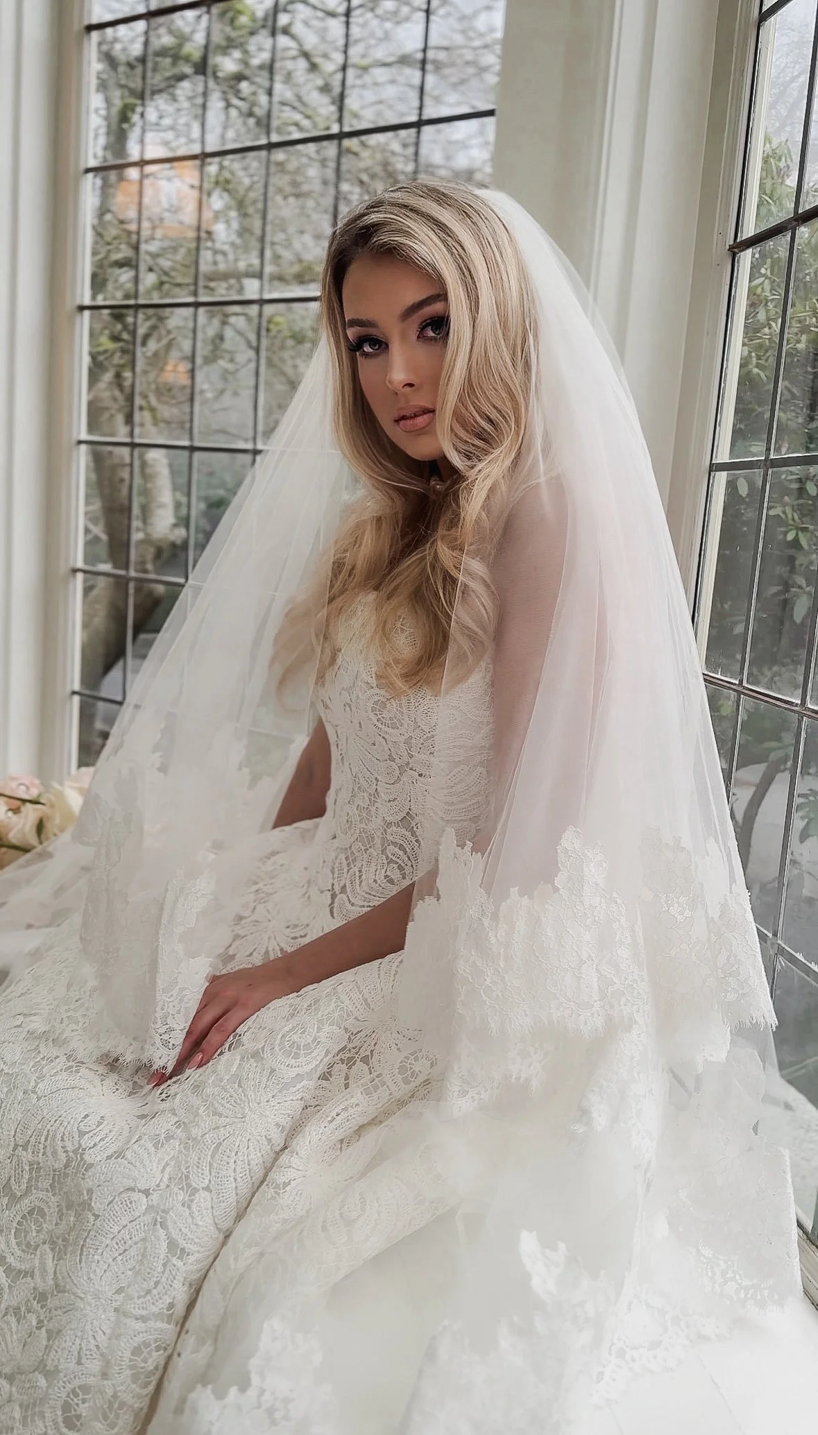 A woman in a wedding dress and veil sitting by a window.