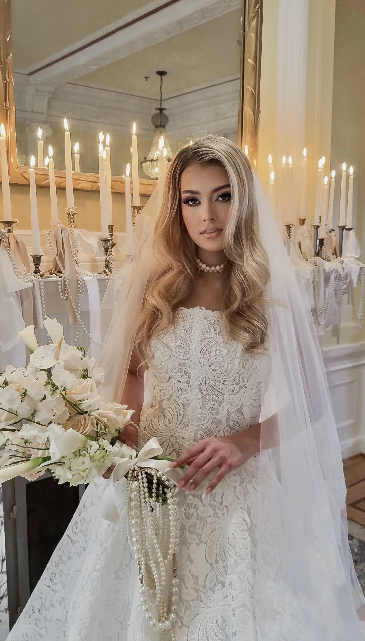 A bride in a white lace wedding dress holding a bouquet of white flowers with pearl necklaces. She is standing in front of a decorated background with lit candles and a large mirror.