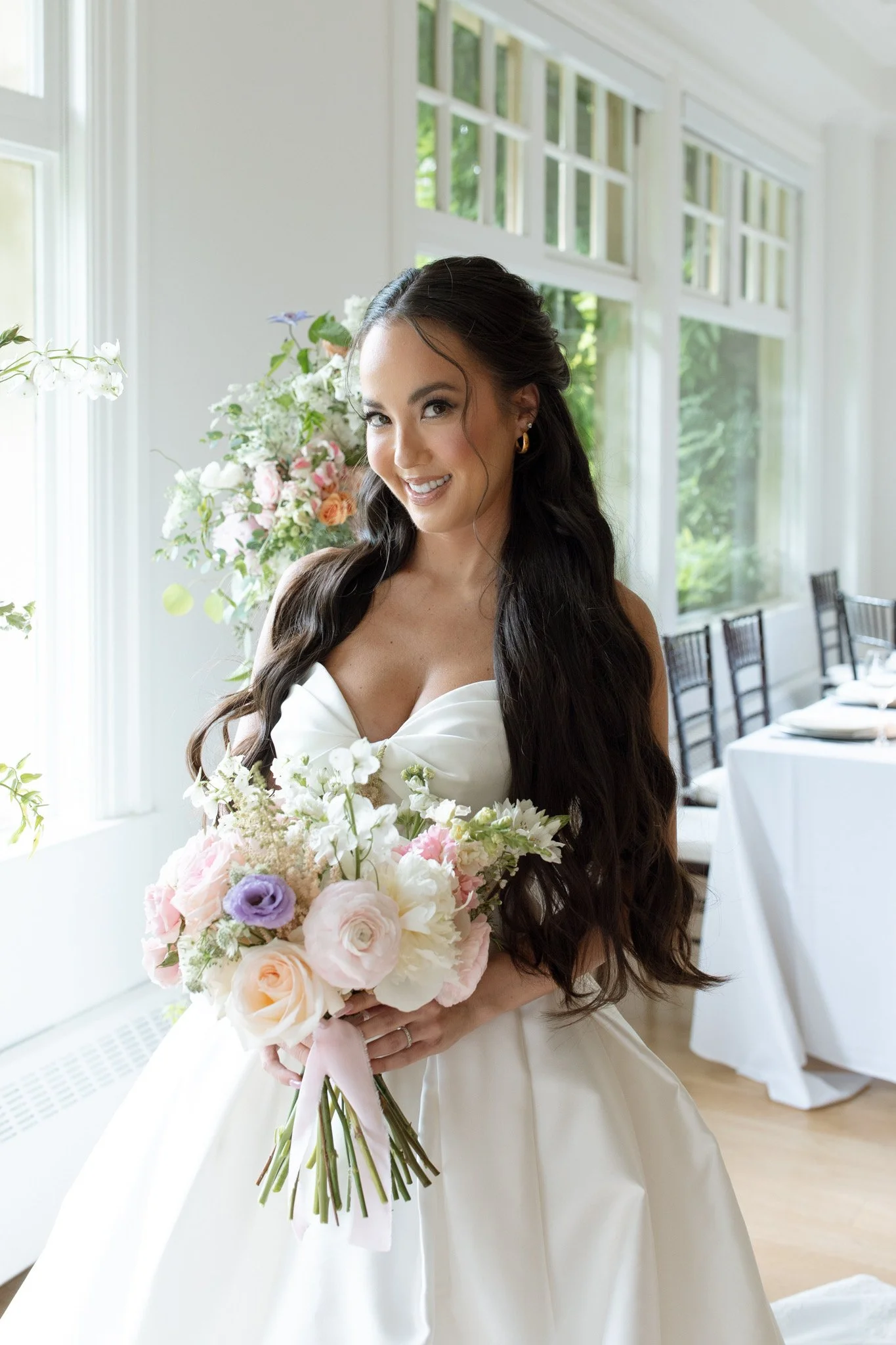A smiling bride with long dark hair in a white wedding dress holding a bouquet of pastel flowers inside a bright room with large windows and floral arrangements.