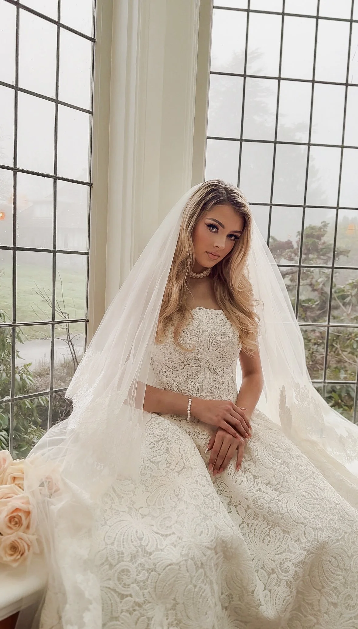 A woman in a wedding dress sitting by a large window with a lace dress and veil.