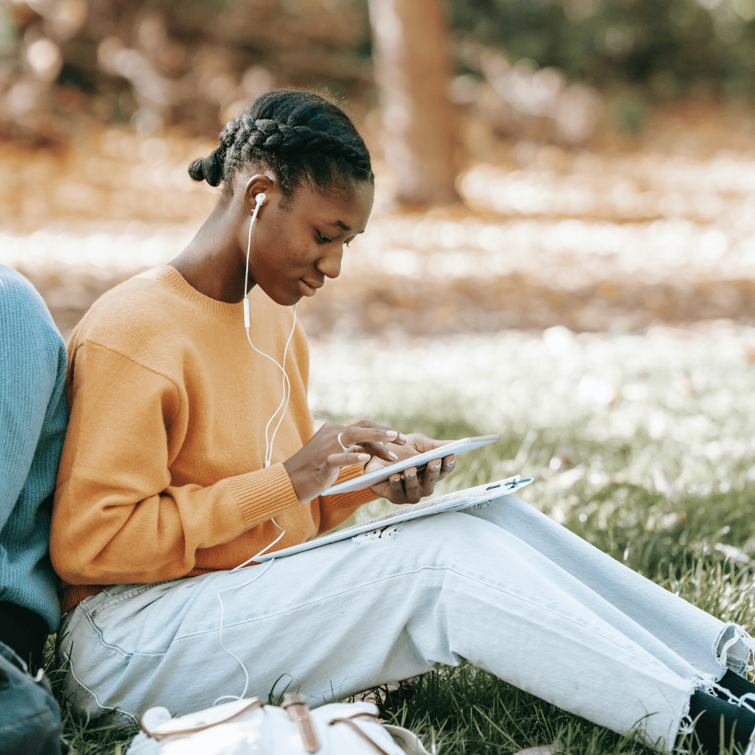A black woman with braids sitting outside listening to music and playing on her tablet device.