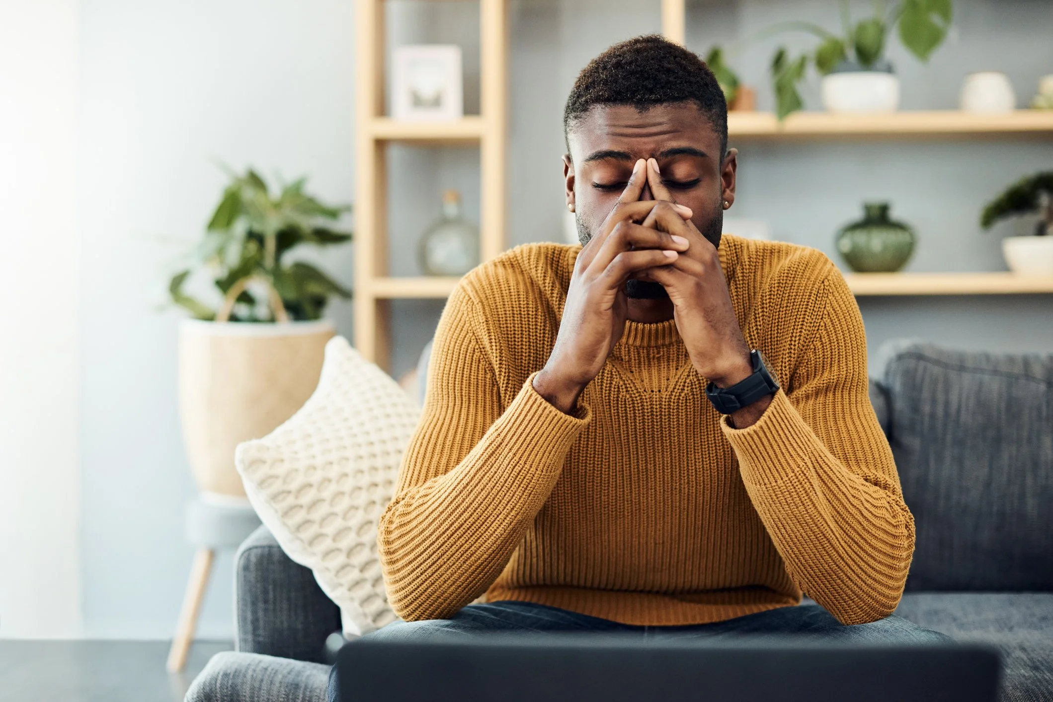 A man sitting on a sofa with closed eyes, appearing contemplative, wearing a mustard-colored sweater in a room with plants and shelves in the background.