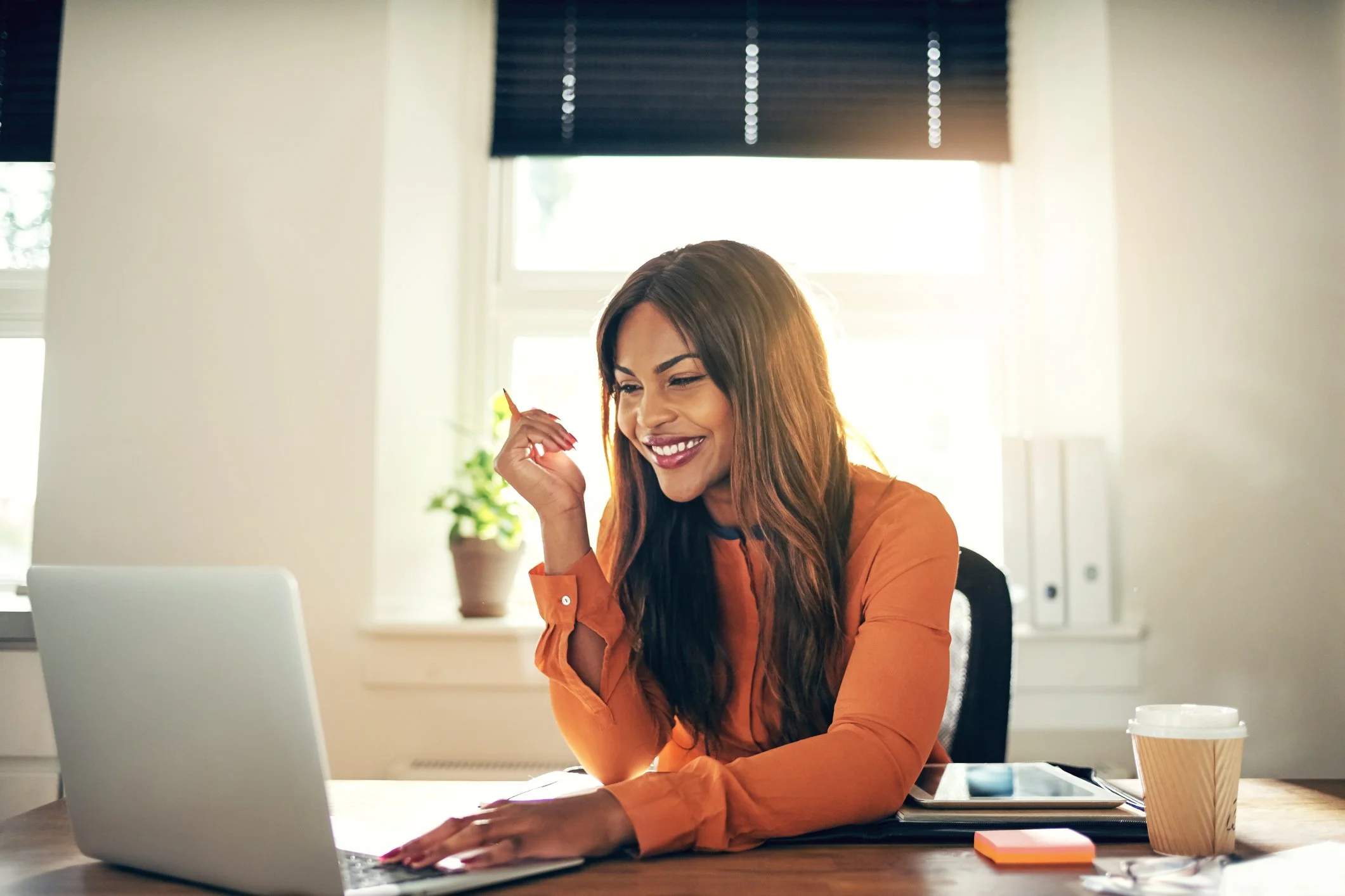 Smiling woman in orange shirt using a laptop at a desk with coffee cup