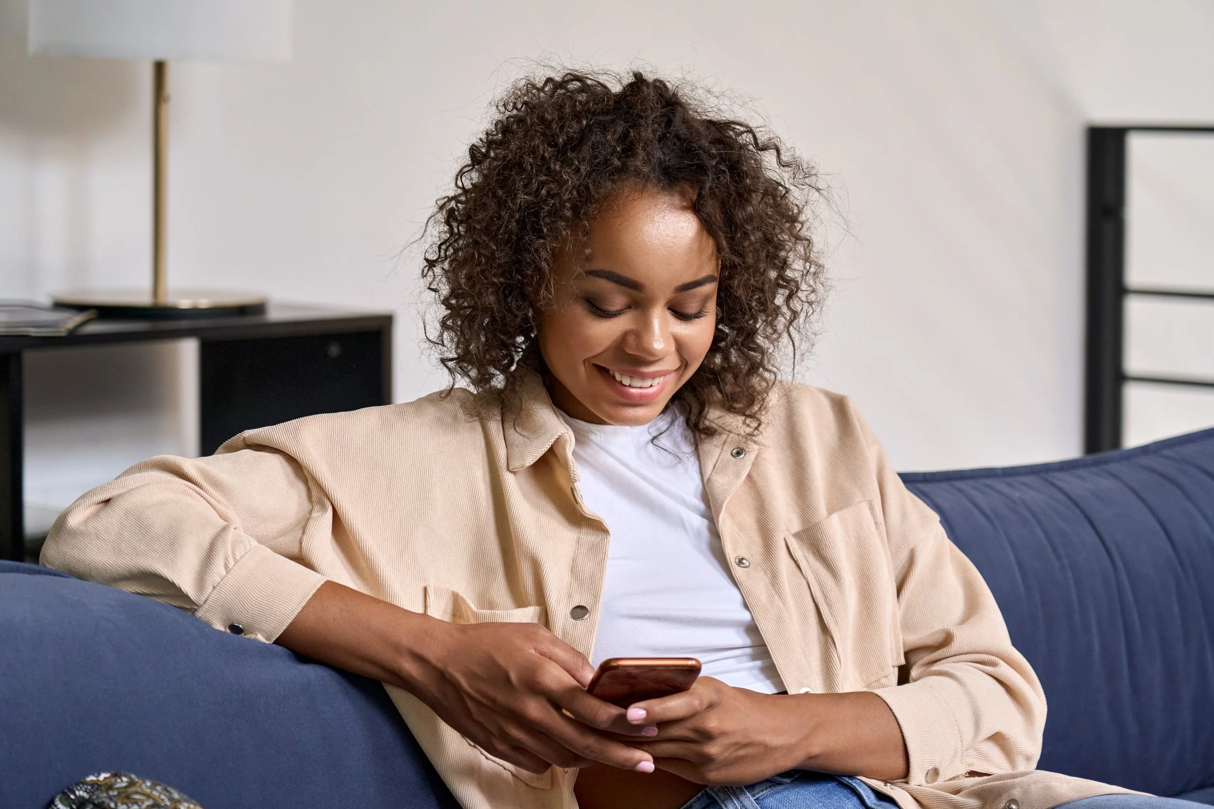 Woman sitting on a couch using a smartphone, smiling, indoor setting.