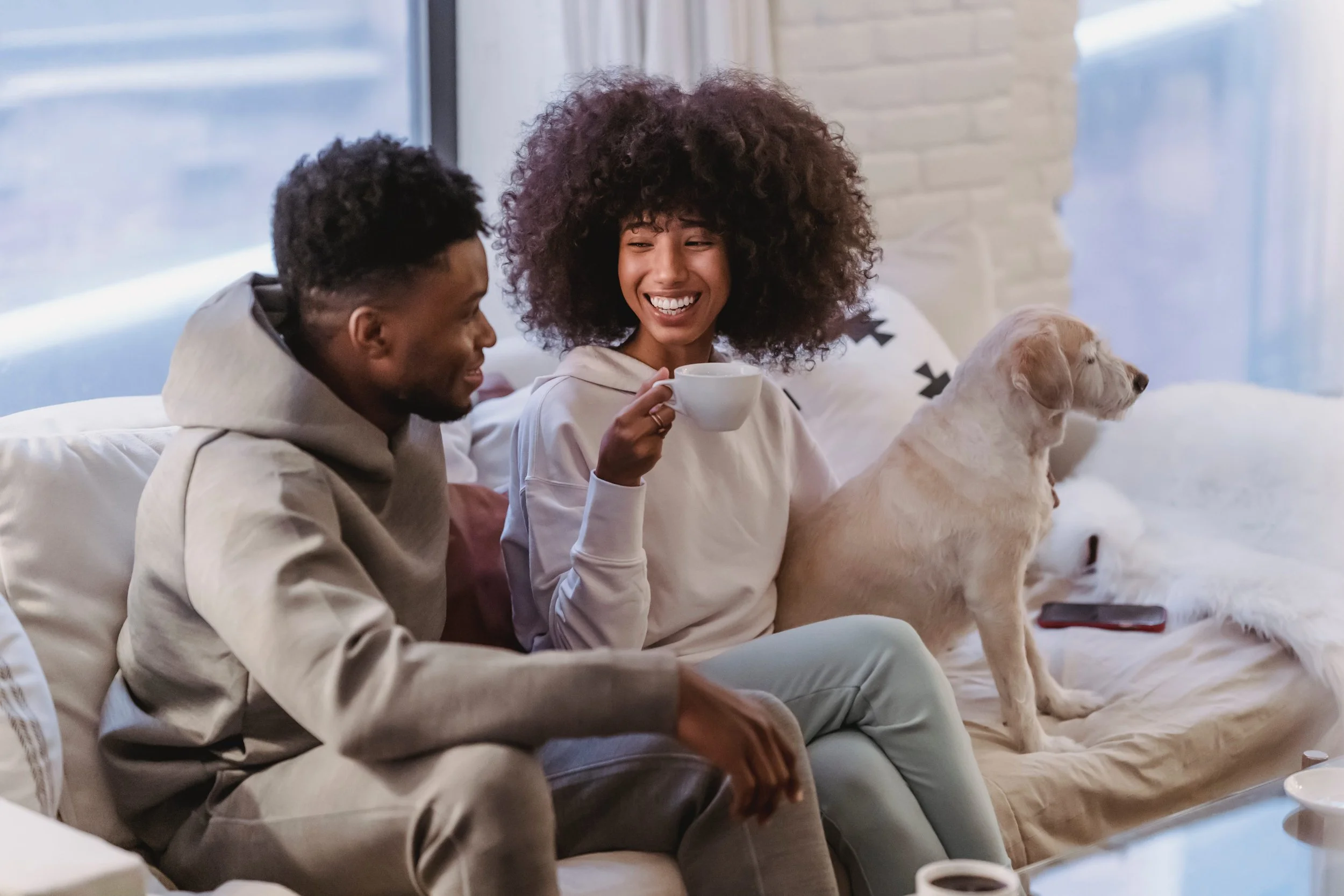 A black couple sitting on the couch and laughing with their dog.