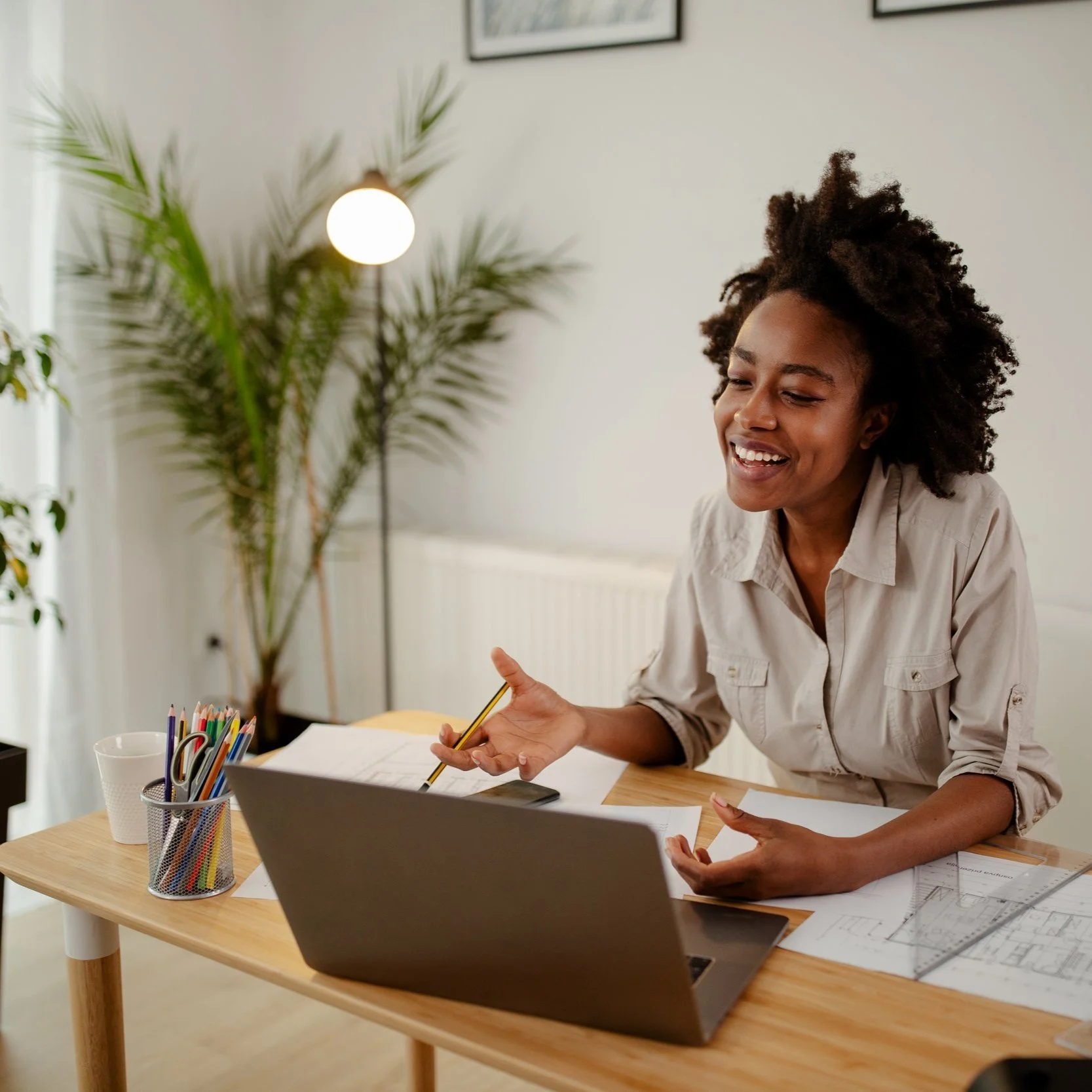 A woman with curly hair smiling while sitting at a wooden desk with a laptop. She is holding a pen and appears engaged in a conversation. The desk also has colored pencils, papers with diagrams, and a smartphone. A potted plant and a lamp are in the background.