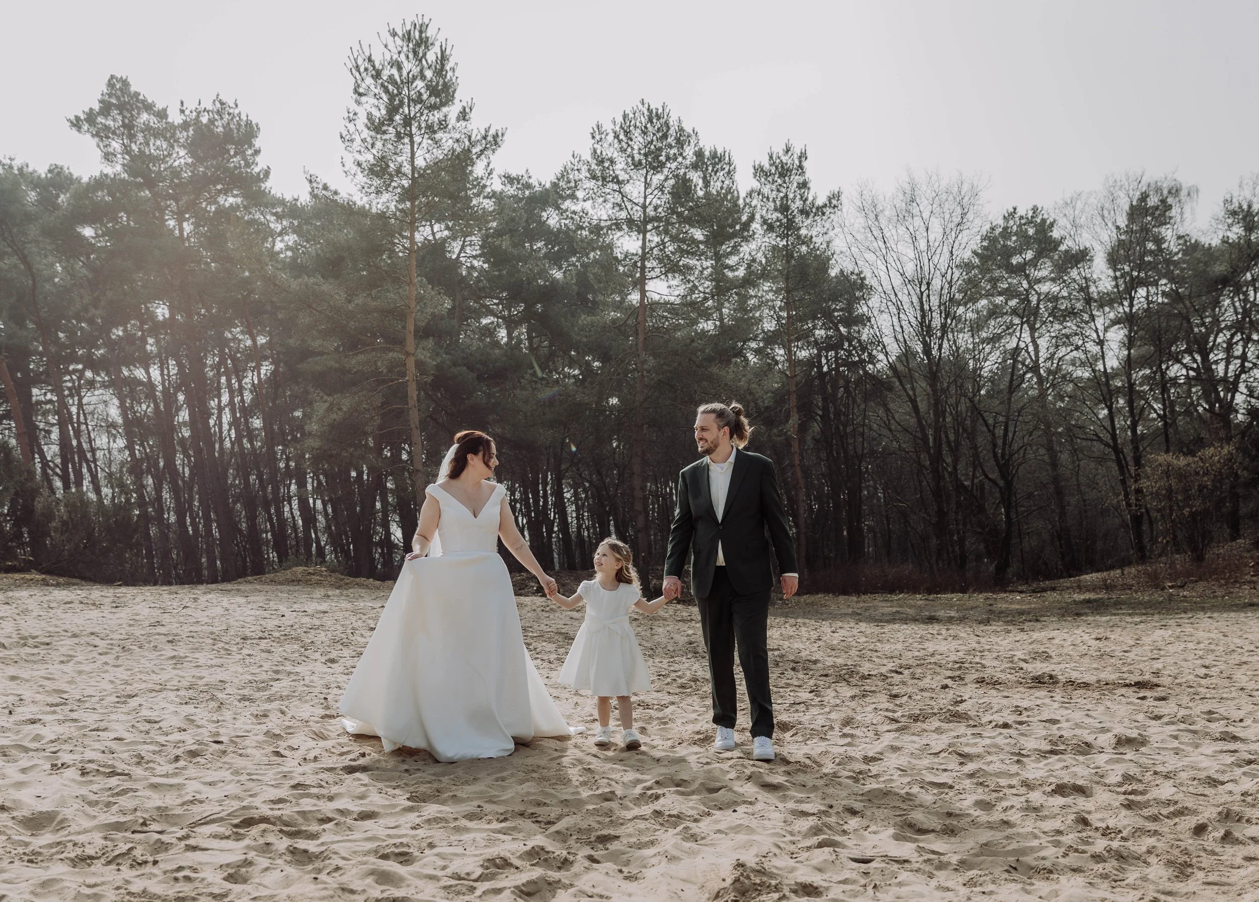 Een bruid en bruidegom lopen samen hand in hand met een jonge meisje op zand omgeven door bomen.