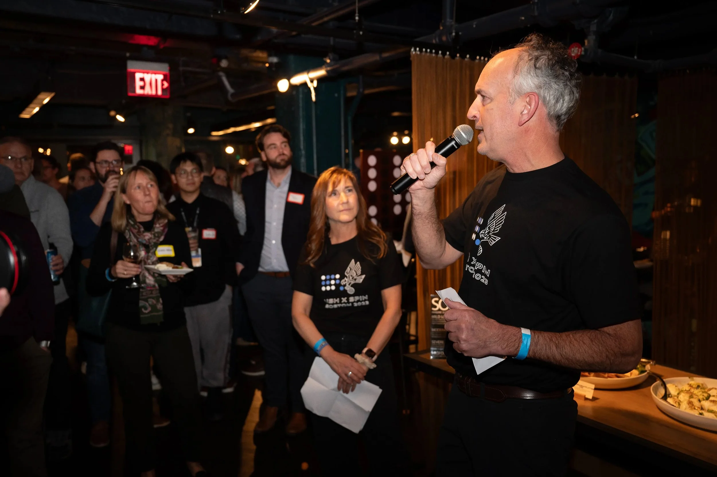 A man, David Corderman stands in the foreground and speaks into a microphone. Founder of the Usher Syndrome Society, Nancy is to his right and a crowd of people watch from nearby. David and Nancy are wearing custom shirts for the Boston SPIN event.