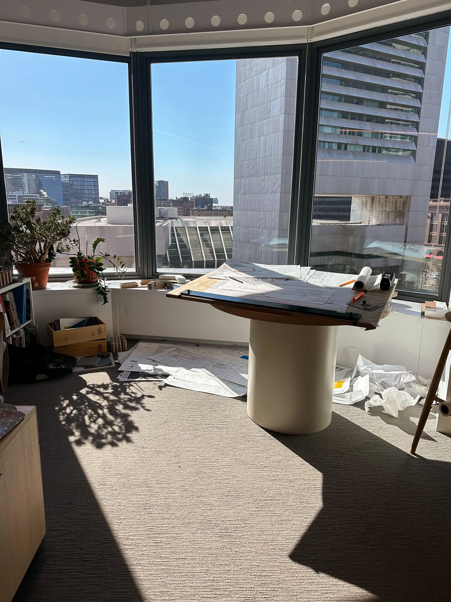 Light streams in onto a set of drawings propped up on a table. There are plants to the left and crumpled paper on the floor. The Boston skyline is visible through the window.