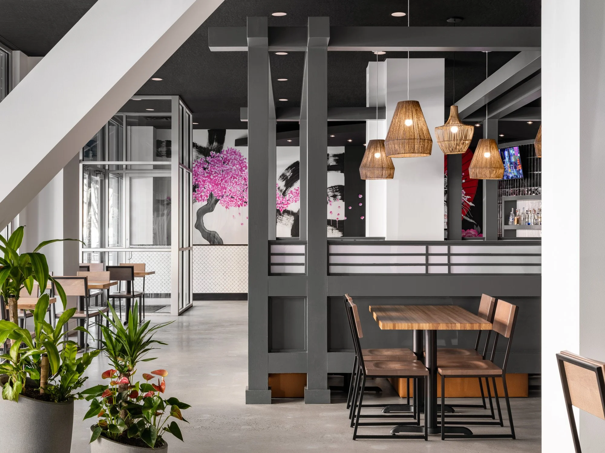 A restaurant interior. The entrance to the restaurant is glass and there are a wooden tables with black accents. There are handing wicker lights above one of the tables. In the background there is a Japanese style painted mural on the wall.