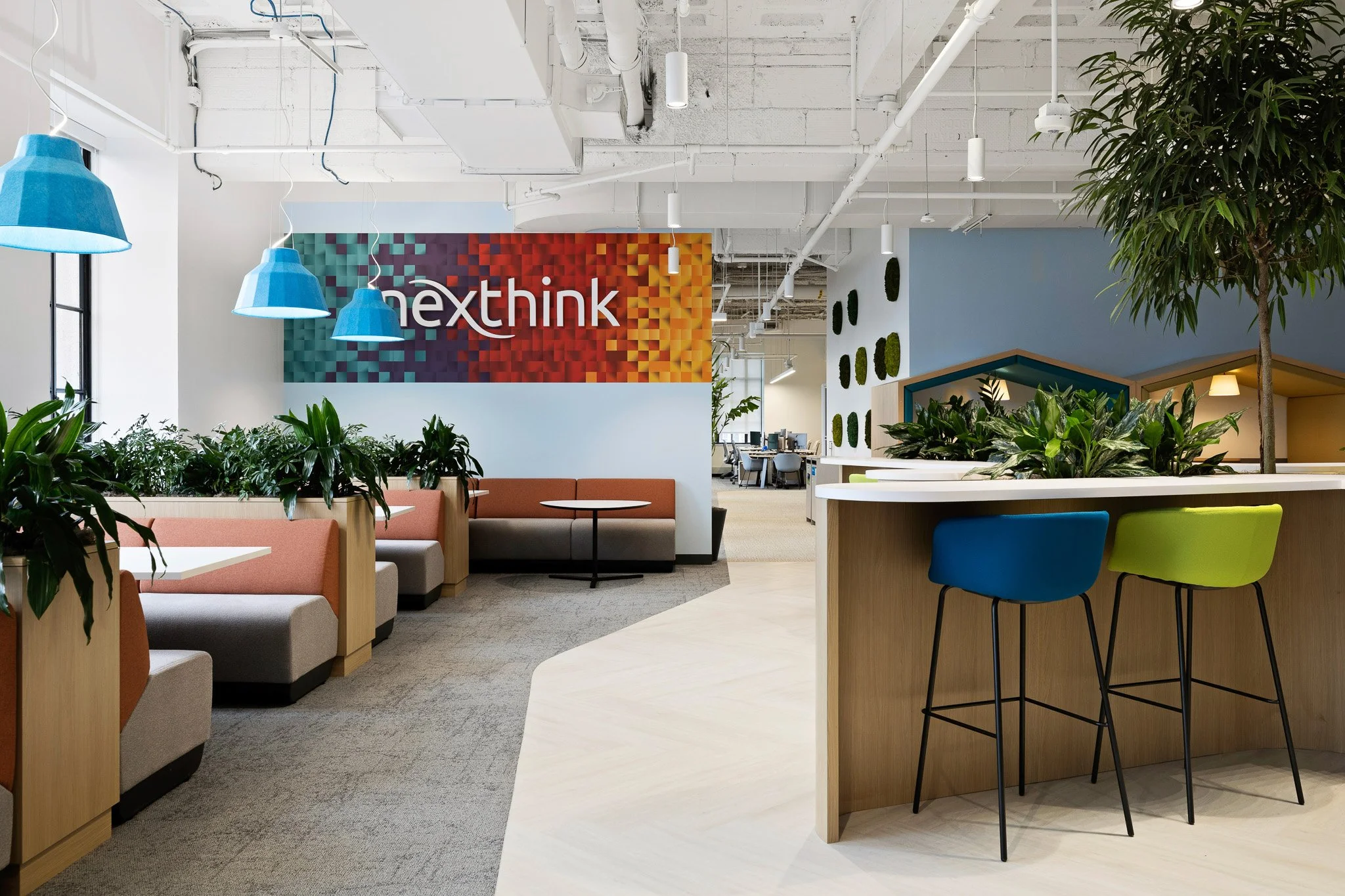 Photo of vibrant corporate interior space with booth style seating on the left, barstool seating on the right, the nexthink logo is colorful on the wall. green plants are placed throughout the space.