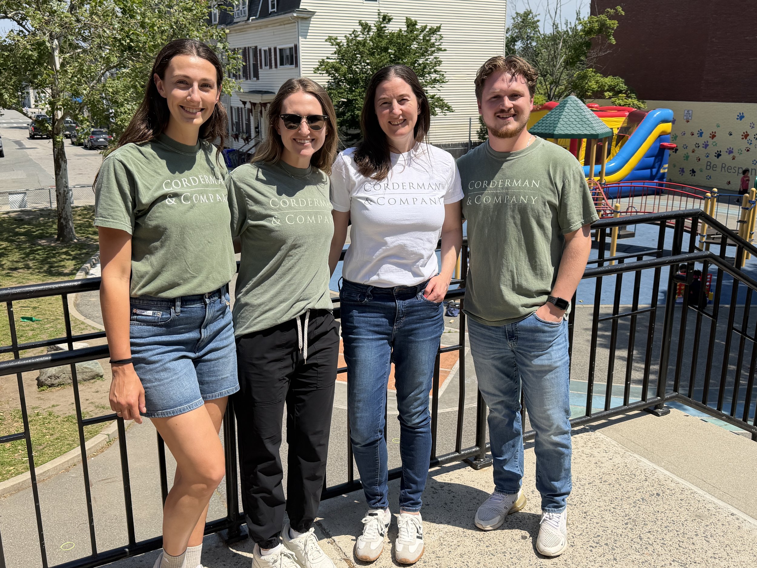Four Corderman & Company team members stand smiling together smiling at the camera. They are wearing Corderman t-shirts and stand at the top of a stairway overlooking a playground at Tynan Elementary School.
