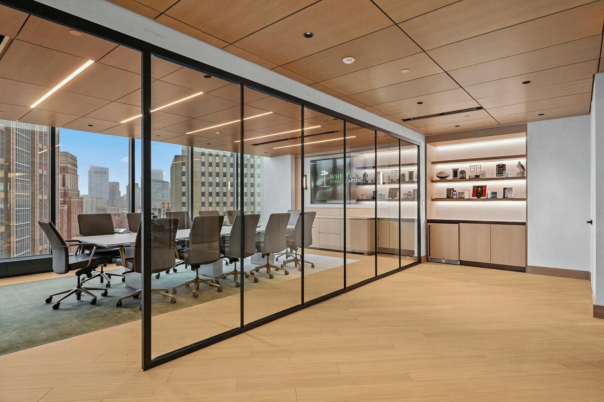 A conference room with a large table and 12 chairs, there are glass office fronts on one side and floor to ceiling windows on the other. A tv displays Wheelock Street Capital's logo.