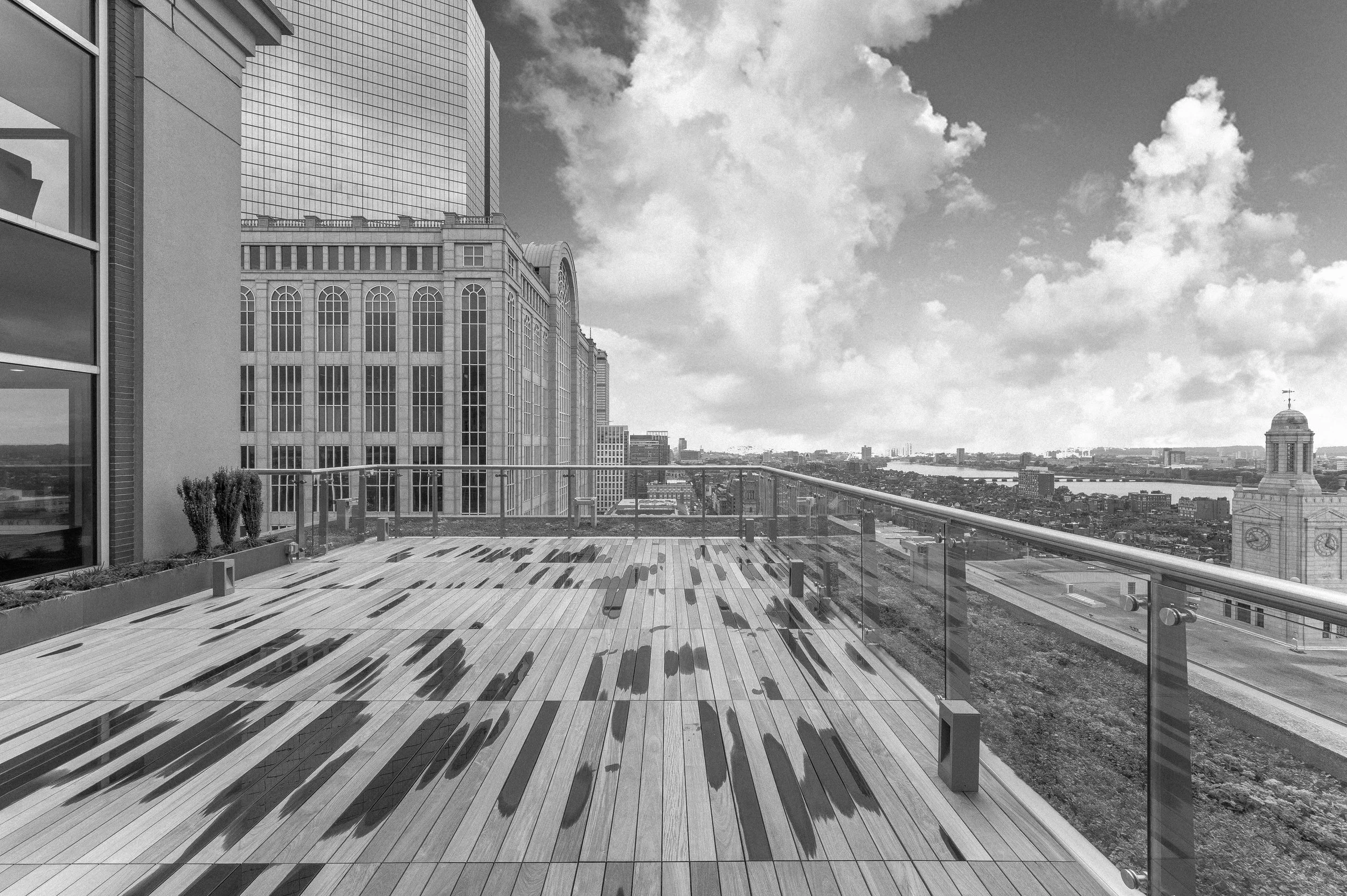 A black and white photo of a roof deck. It has just rained so some spots are a little wet, there is a glass and metal railing. 500 Boylston and Boston's Back Bay skyline are a captivating backdrop.