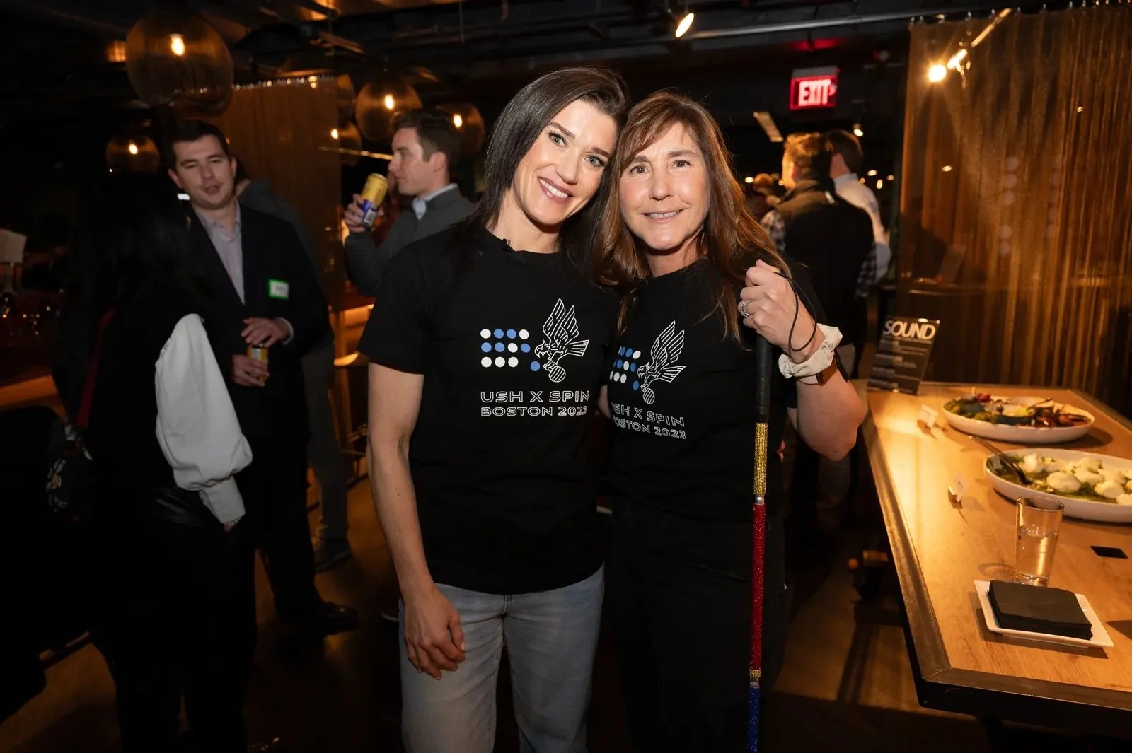 Two women wearing black shirts stand together smiling at the camera. One woman holds a bedazzled cane. They are wearing custom shirts for the SPIN Boston event.