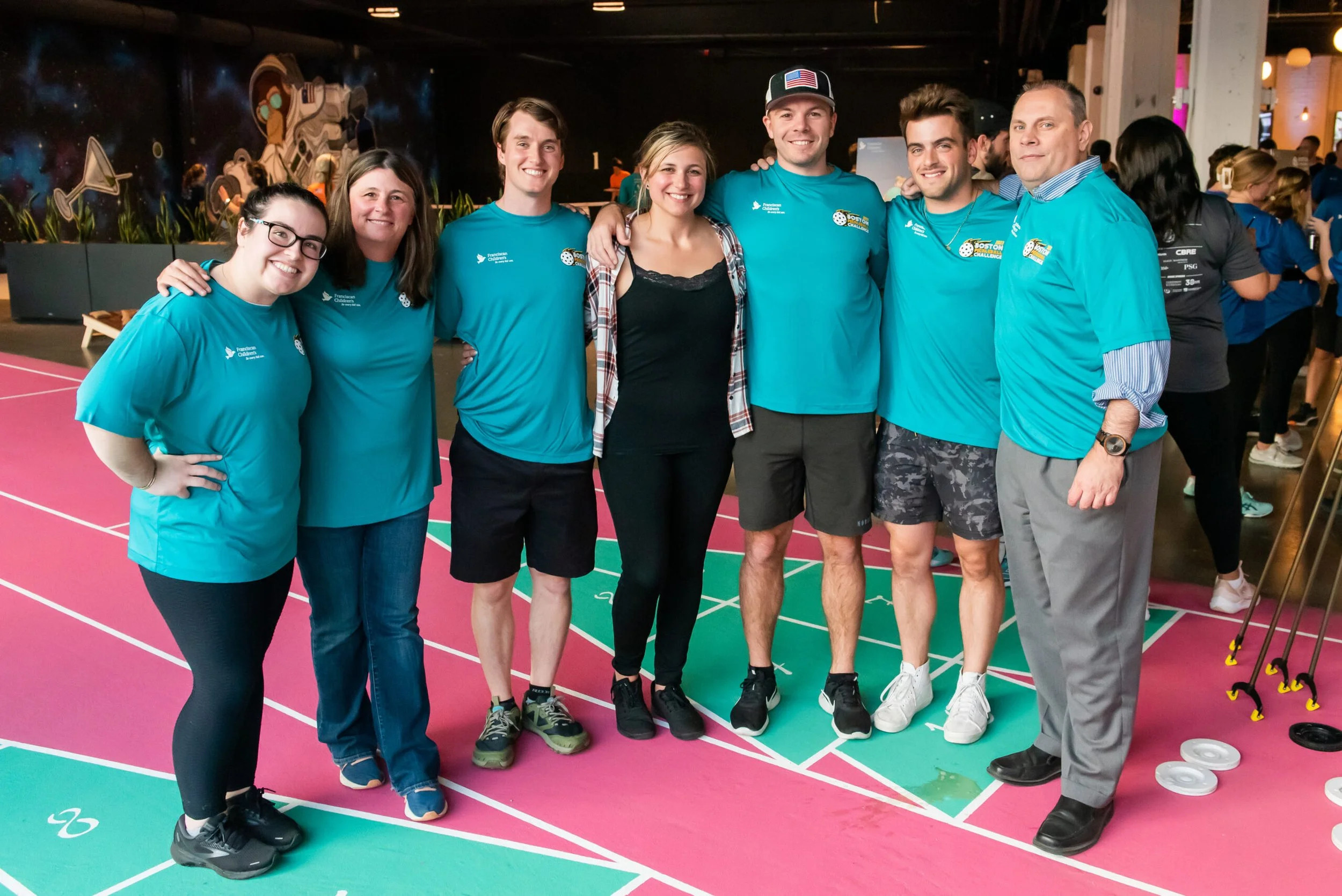 A group of Corderman Employees posing at a Charity Pickleball event. They are wearing matching teal event t-shirts standing on a shuffleboard court