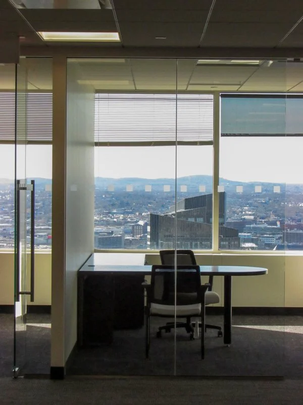 An office space with glass partitions and a view of the city and surrounding land. There is an empty desk and two chairs, the blinds in the window cover part of the view.