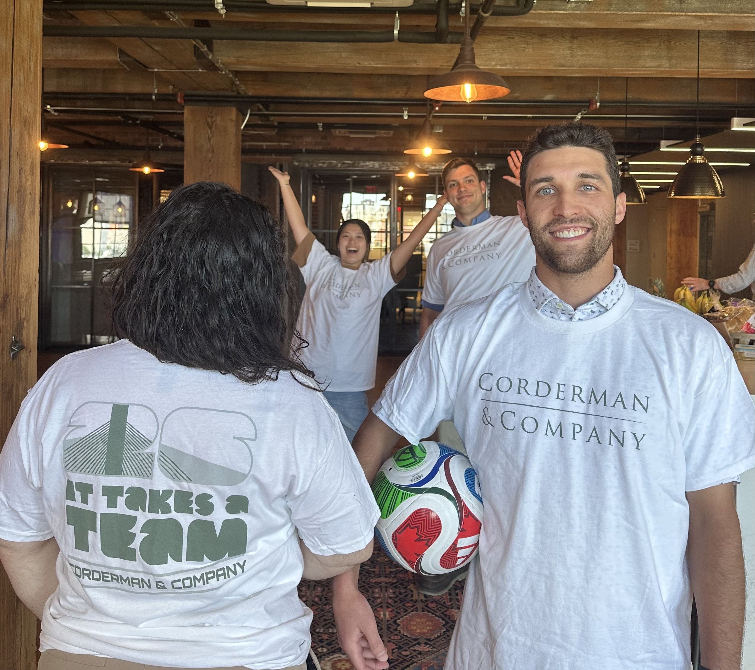 Four Corderman employees are in the frame. One has her back to the camera but on the back of her shirt is a large 26 and the words "It Takes a Team" and the Corderman name. The man next to her smiles at the camera and holds a soccer ball.