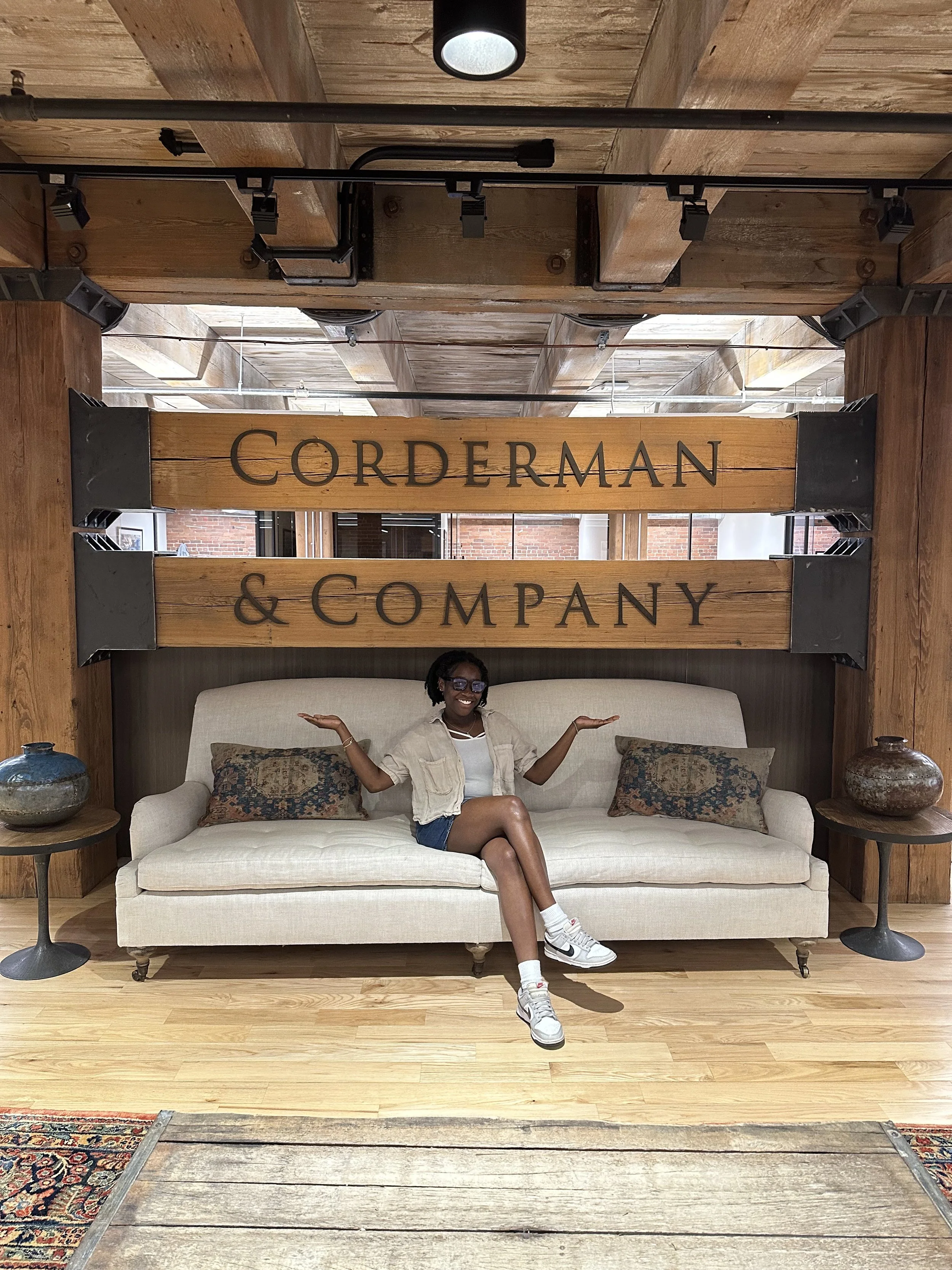 A young woman sits on a couch underneath a sign that reads Corderman & Company across two large wooden beams. She is crossing her legs, smiling, and holding her hands out posing for the camera. She joins the team as a summer Accounting intern.