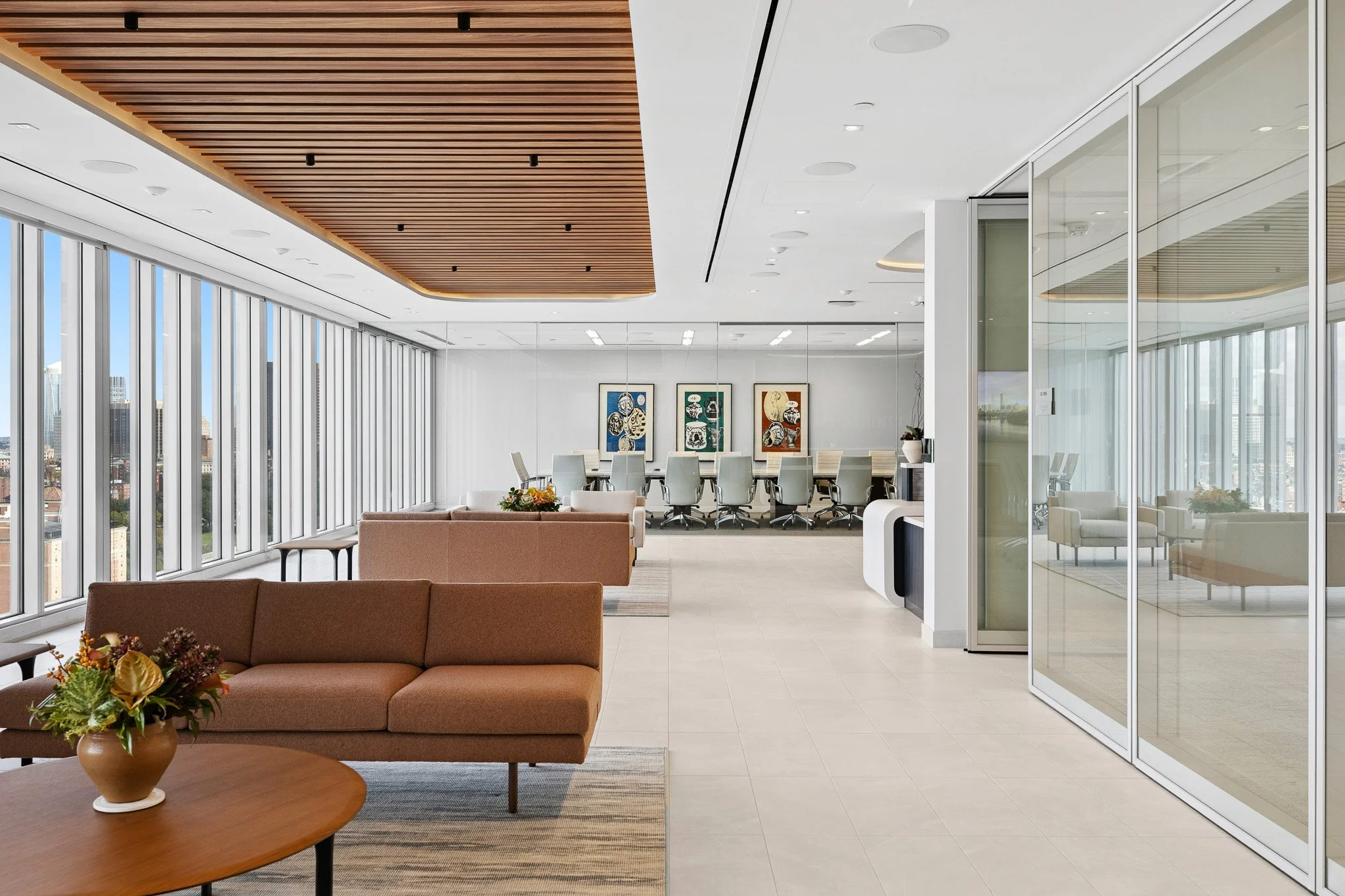 An interior space built by Corderman with offices lining the right side and a seating area with a bouquet of flowers, a table, sofas, and rugs to the left next to windows overlooking Boston. There is a conference room at the end of the hall.