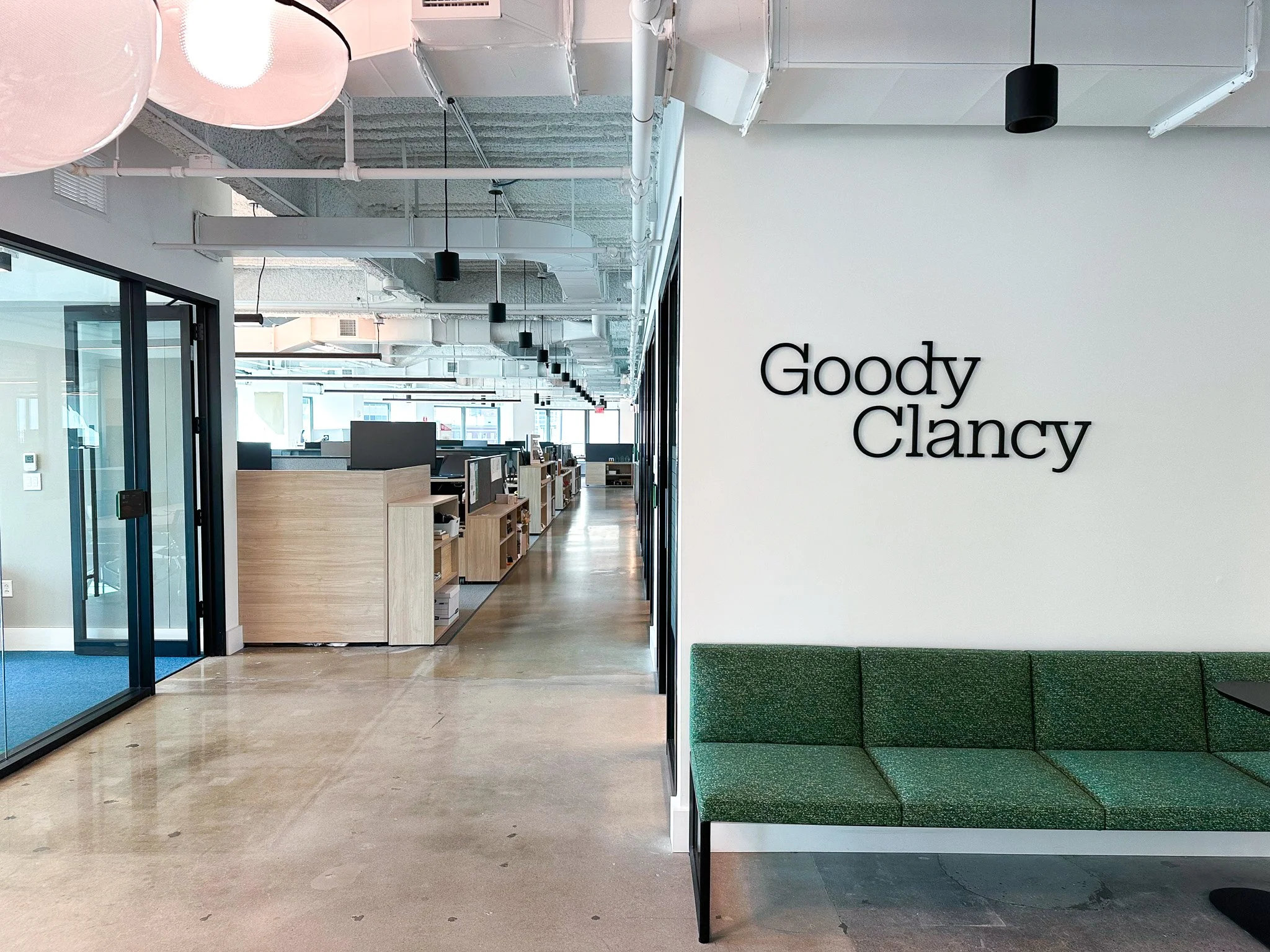 Goody Clancy's logo is displayed on a white wall to the right, above a green couch. A long hallway extends down the left side of the frame and some of the workstations are visible at the design firm's 99 High Street office.
