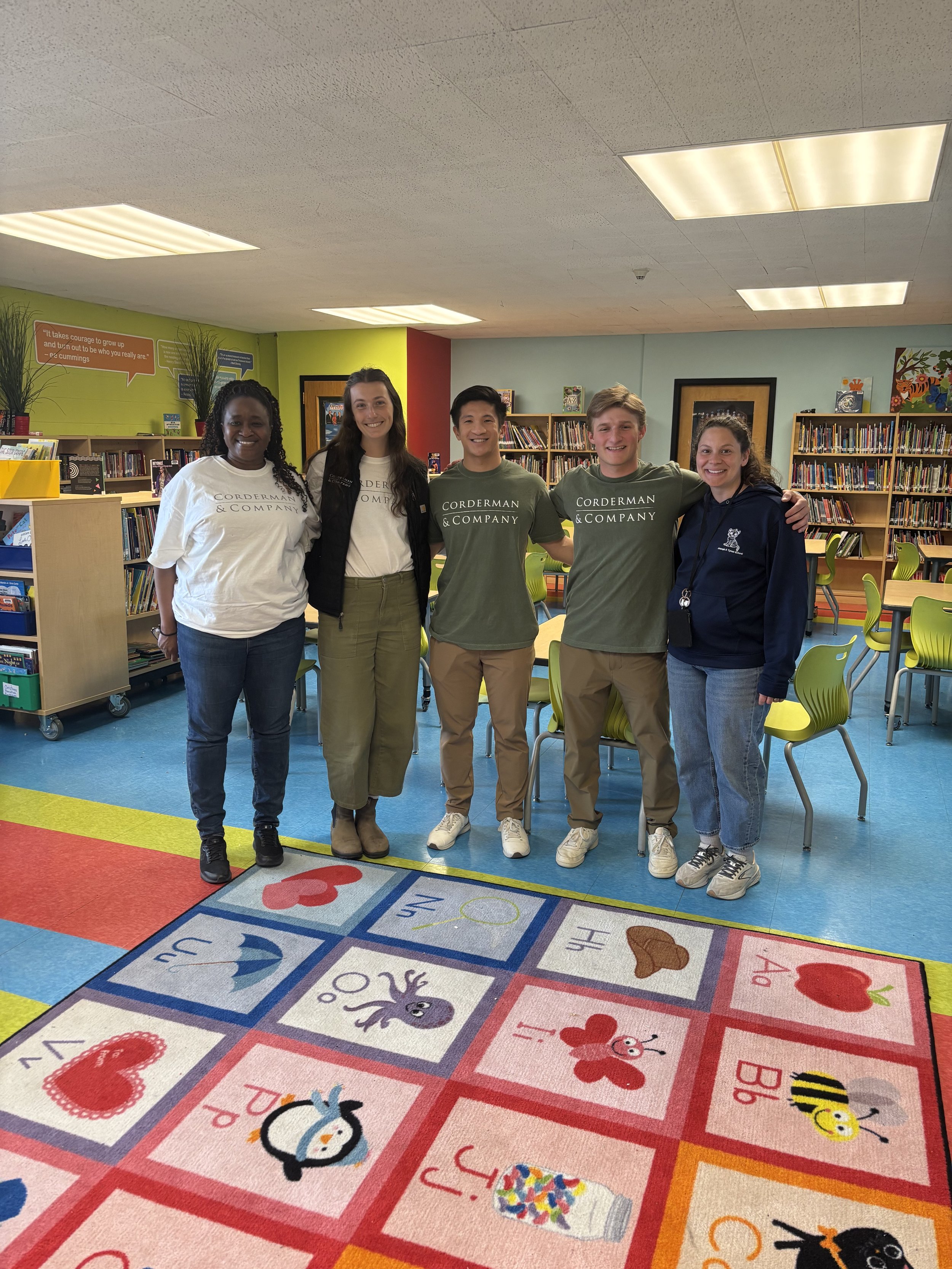 Four Corderman & Company employees and a Tynan Elementary School staff member are standing smiling for the camera. They are standing by a colorful alphabet rug and there are bookshelves and tables behind them.