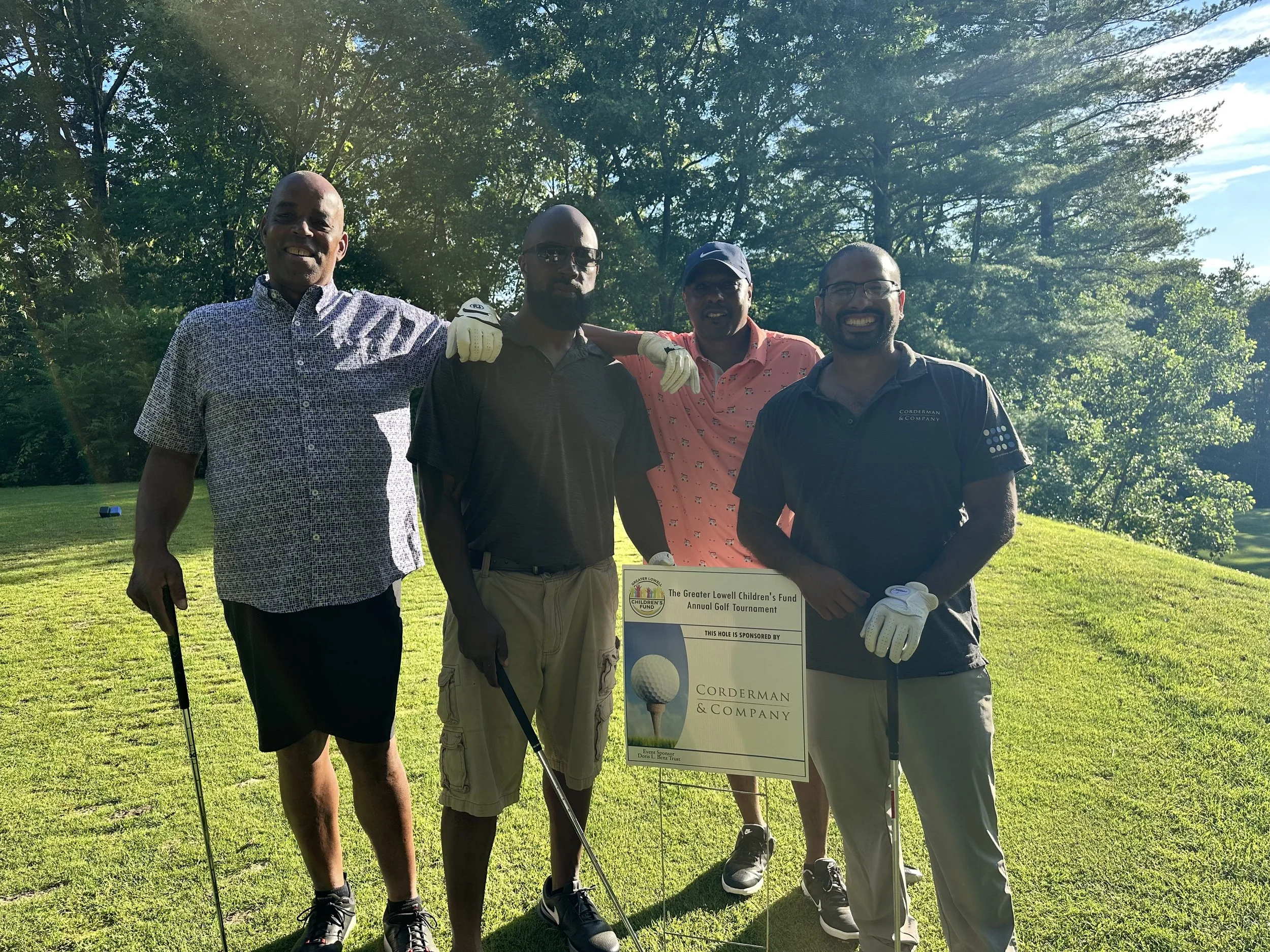 Four men stand posing and smiling at the camera, they are holding golf clubs and standing by a Hole Sponsor sign with the Corderman logo. They are standing on a golf course participating in the Greater Lowell Children's Fund golf tournament.