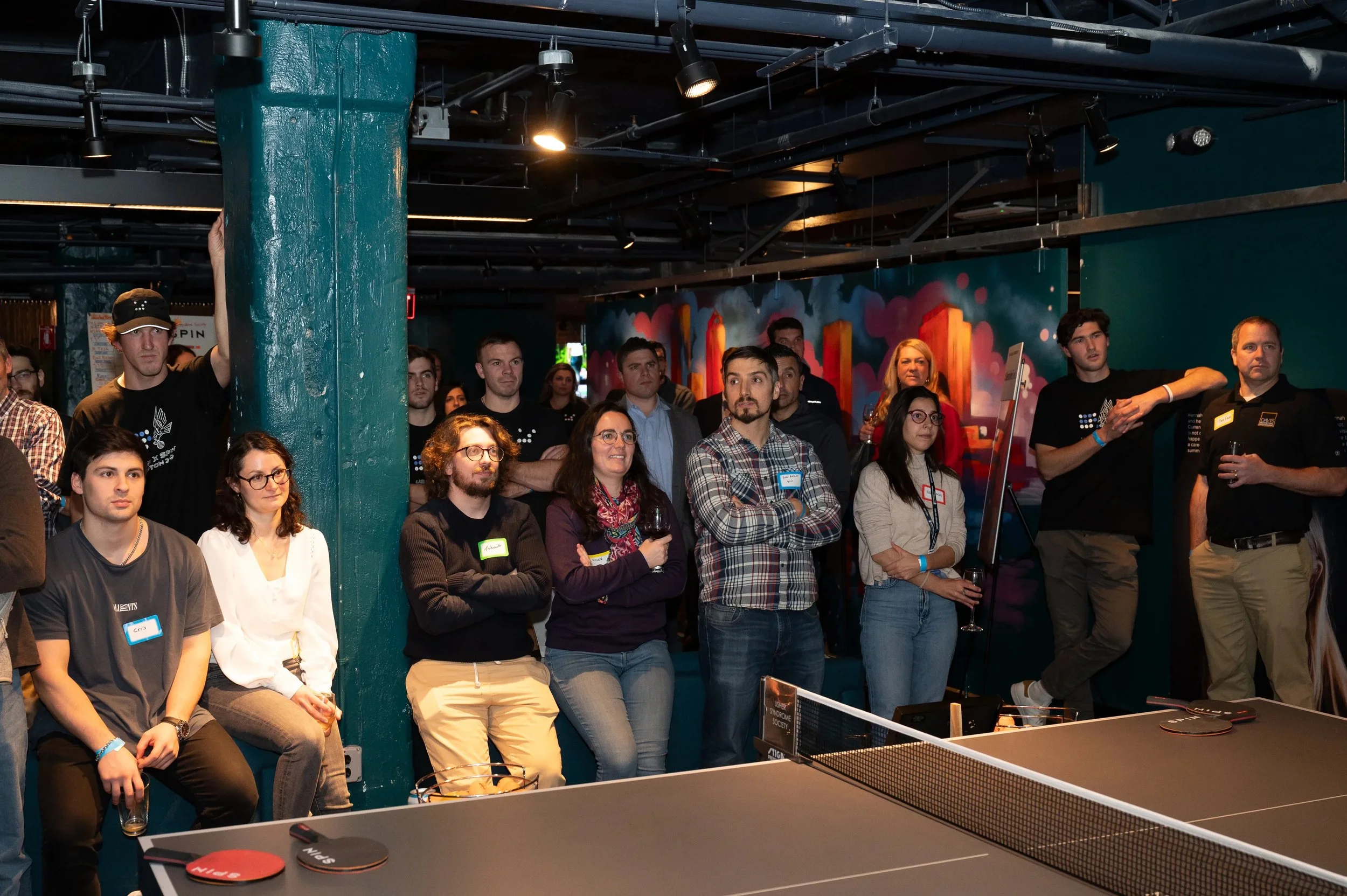 A ping pong table with paddles in the corners is in the foreground. A crowd of people stand and sit on the other side. They all look towards the same area. The group is in attendance at the Ush Society's fundraising SPIN event in Boston.