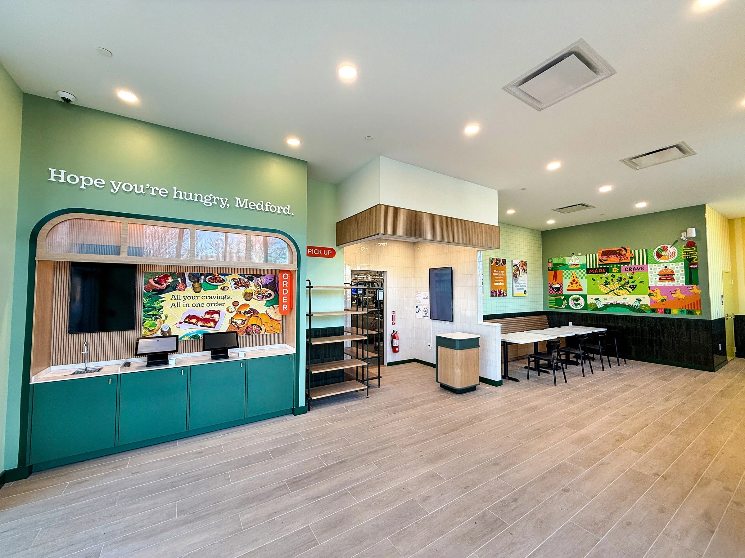 The interior of the new Wonder location at Station Landing in Medford, MA. A kiosk ordering area is on the left, then shelves for take-out orders, a door leading to the commercial kithen, and seating and artwork on the walls to the right.