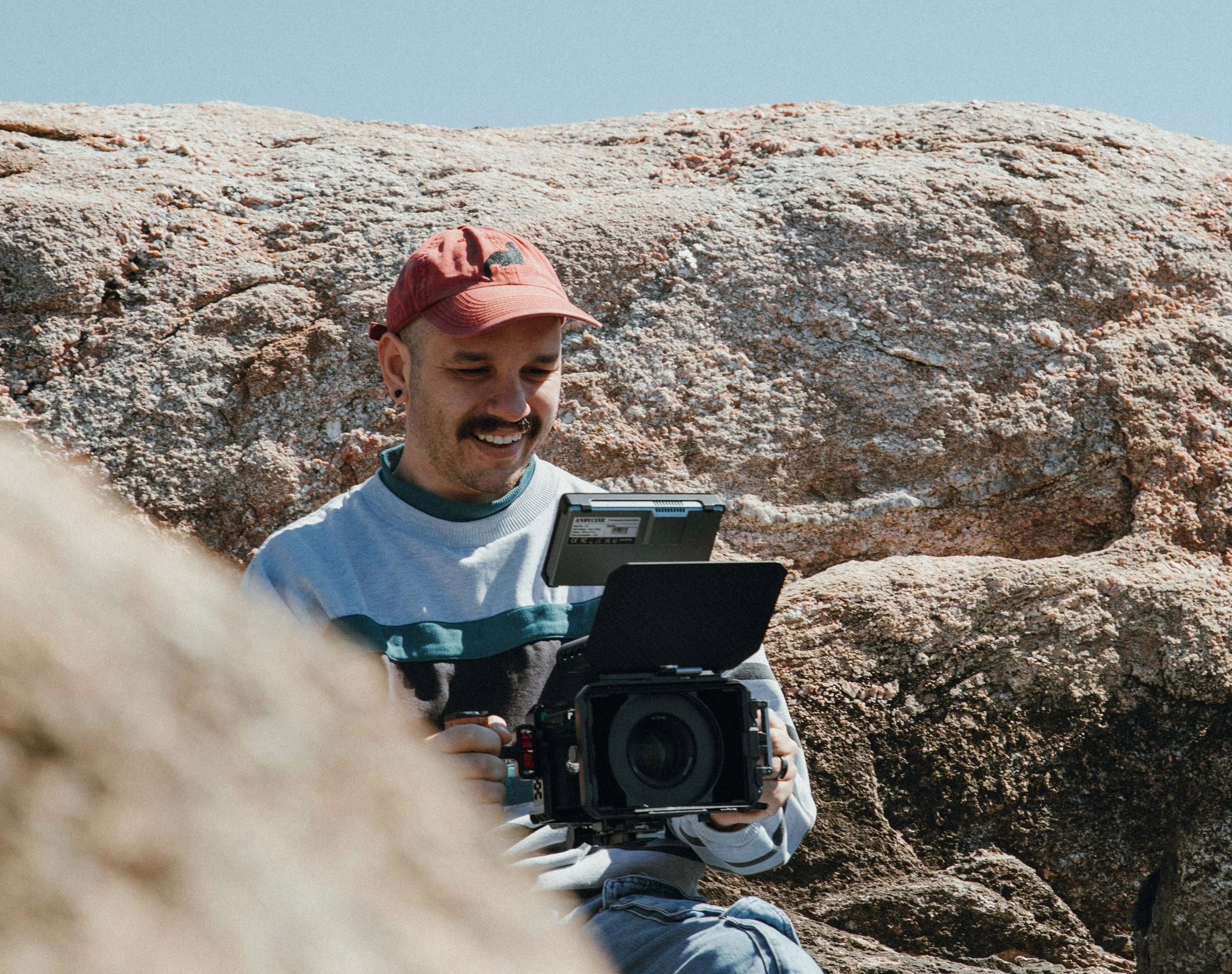 A man with a red cap and a mustache smiling while holding a video camera in front of a large rocky formation.