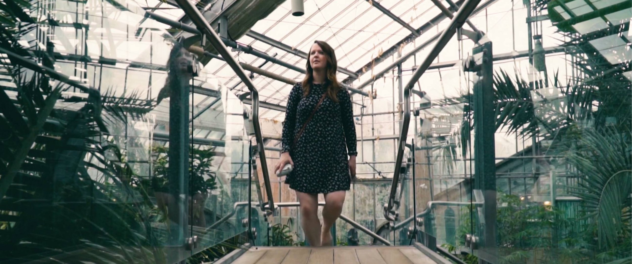A woman in a black floral dress walking through a greenhouse with glass walls and ceiling, surrounded by green plants.