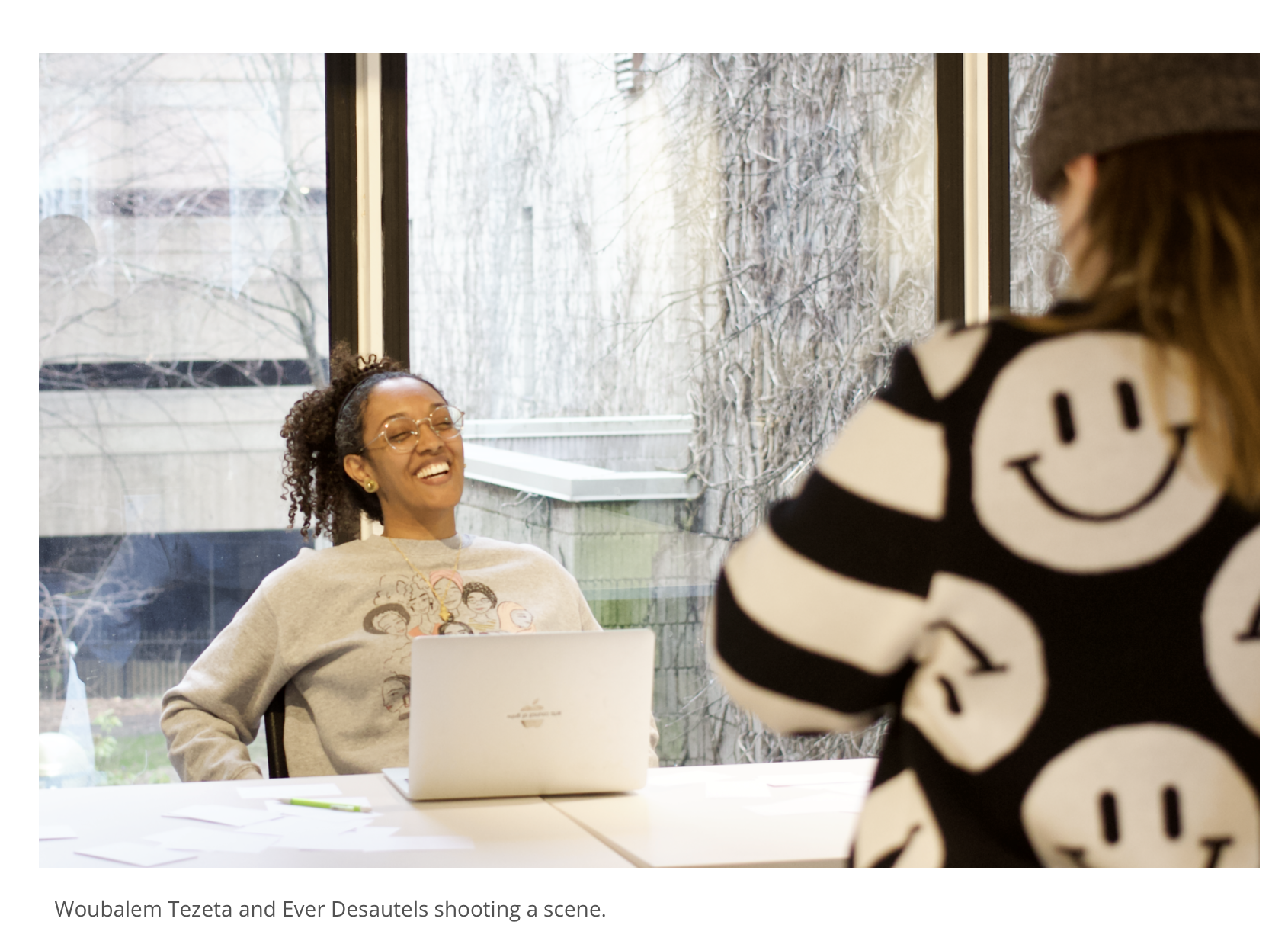 Two women in a meeting room, one sitting and laughing while using a laptop, the other standing with their back to the camera, wearing a black and white striped sweater with smiley face design, during daytime with a snowy outdoor scene visible through large windows.