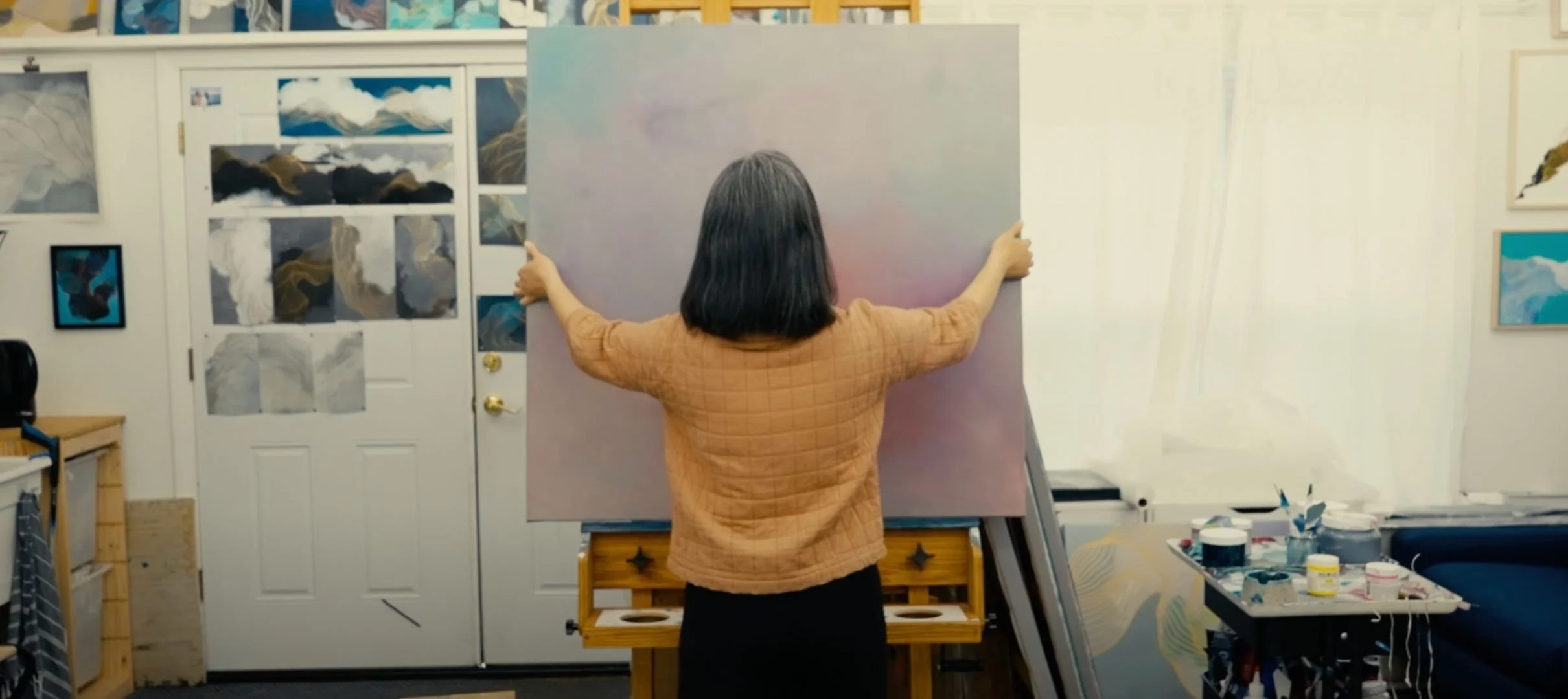 An artist standing in an art studio holding up a large canvas with their back to the camera. The studio has paintings on the walls and art supplies on a cart to the right.