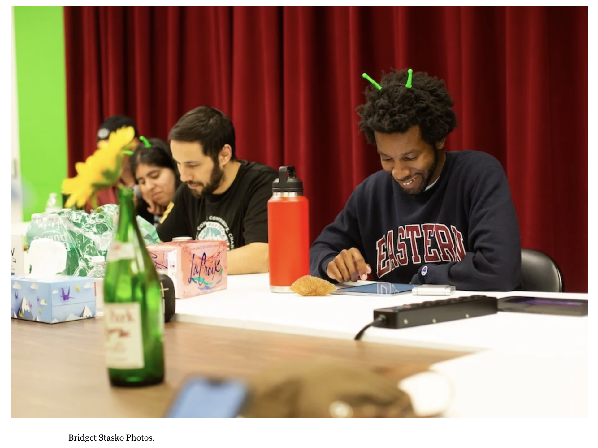 People sitting at a table with red curtains in the background, some wearing green antenna headbands, and various items including a sunflowers, gift boxes, a red water bottle, and a tablet.